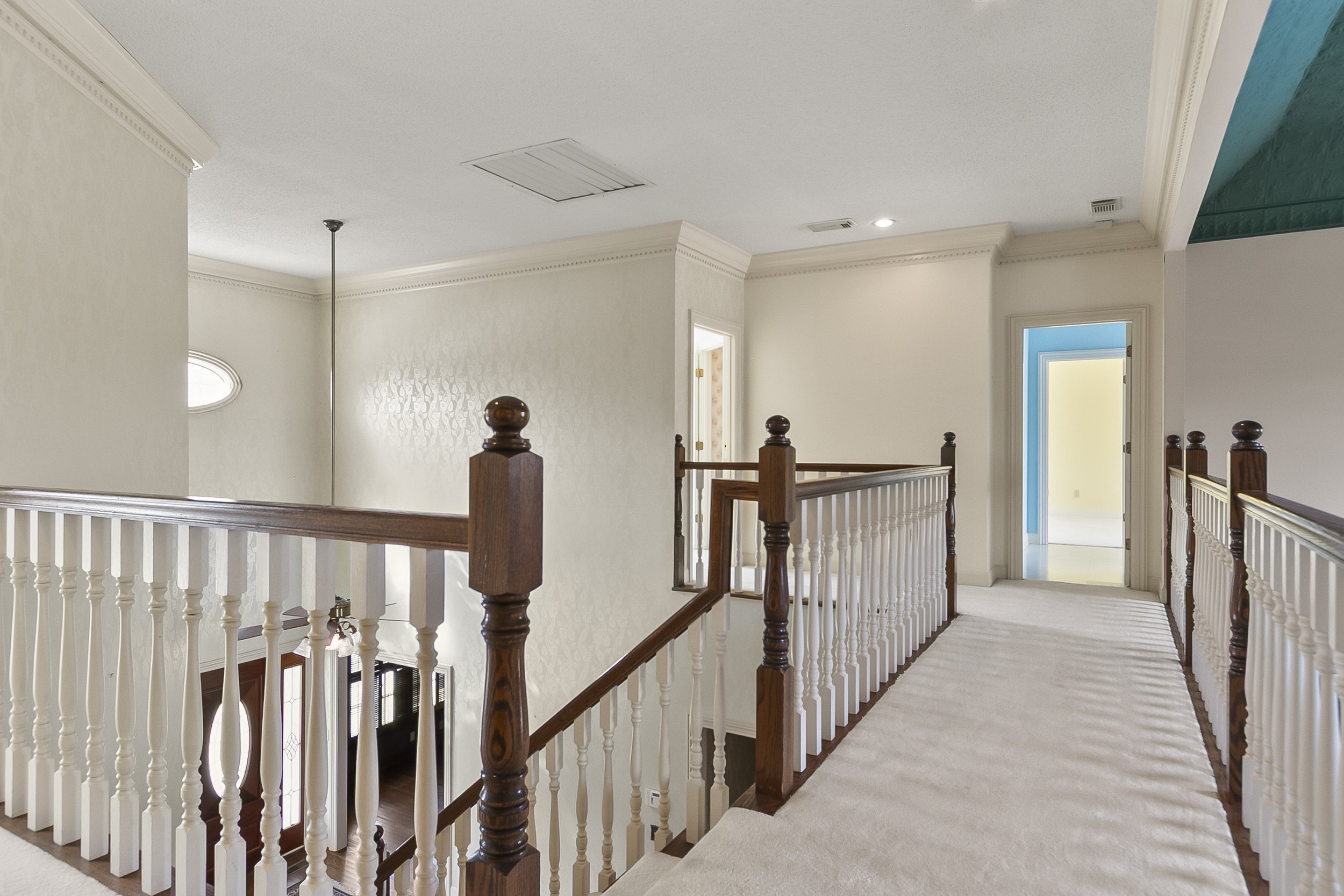 Upper floor hallway with white walls, beige carpet, wooden railings, and multiple doorways, some painted in bright colors.
