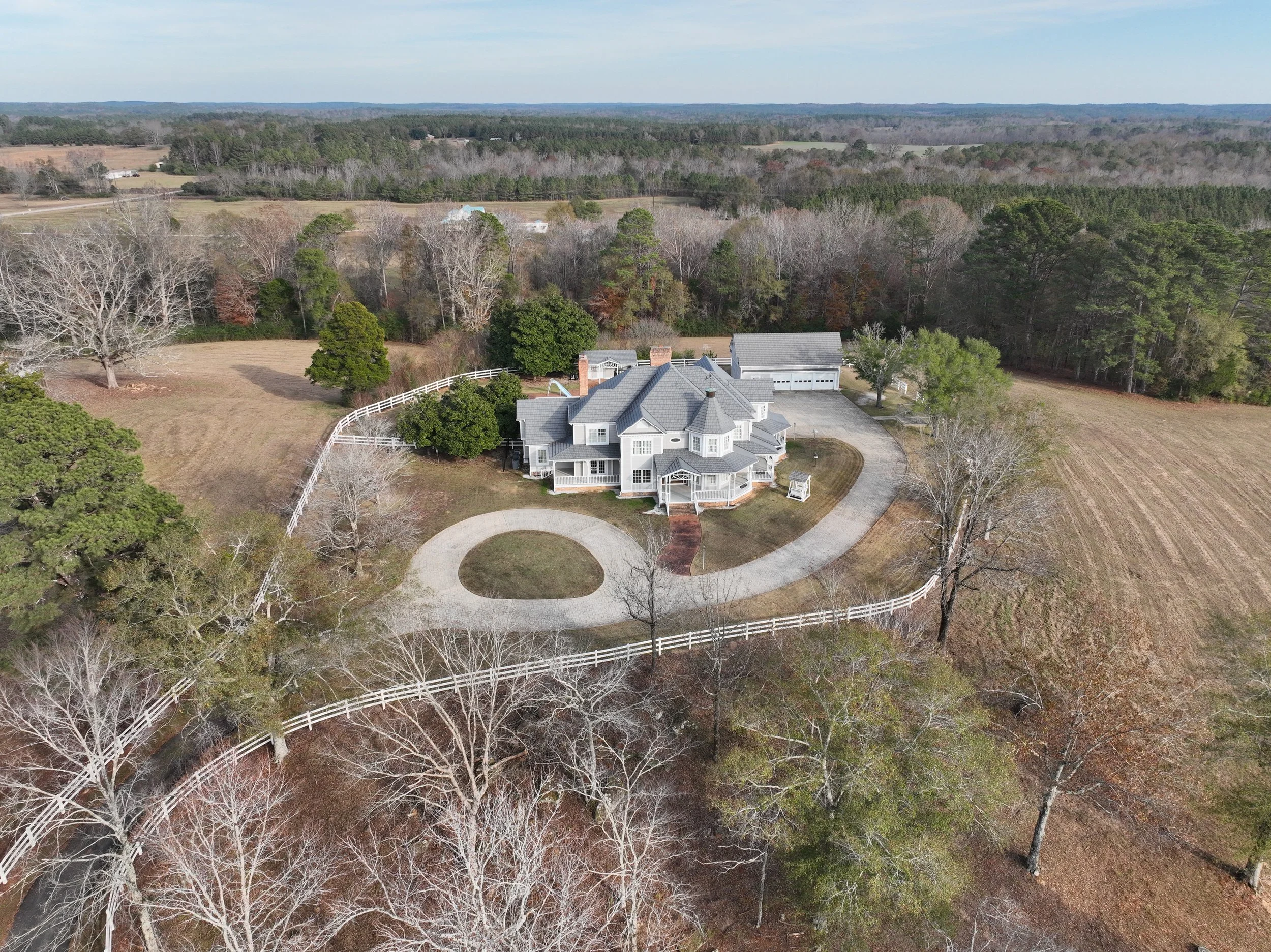 A large house with a gray roof and white exterior surrounded by trees and a white fence, with a circular driveway in front, located in a rural area with fields and forests in the background.