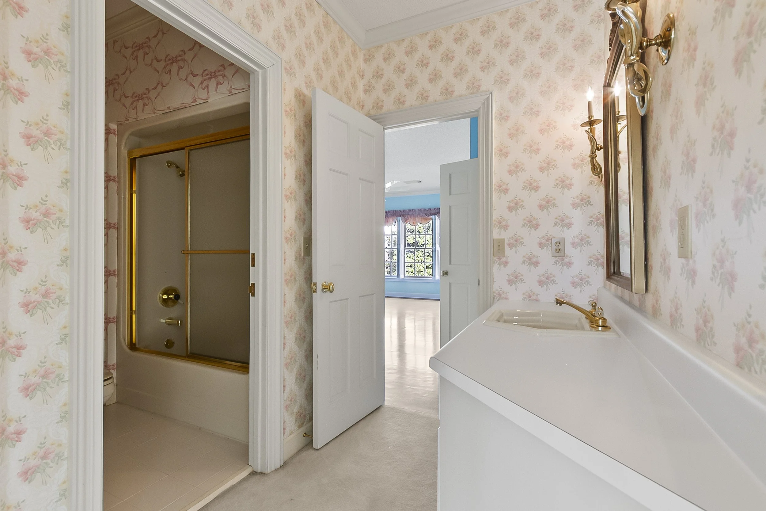 Bathroom with floral wallpaper, gold-framed mirror, and wall-mounted candles. A laundry sink with a gold faucet is visible. A shower with a gold frame and sliding door is in a separate room with floral wallpaper.