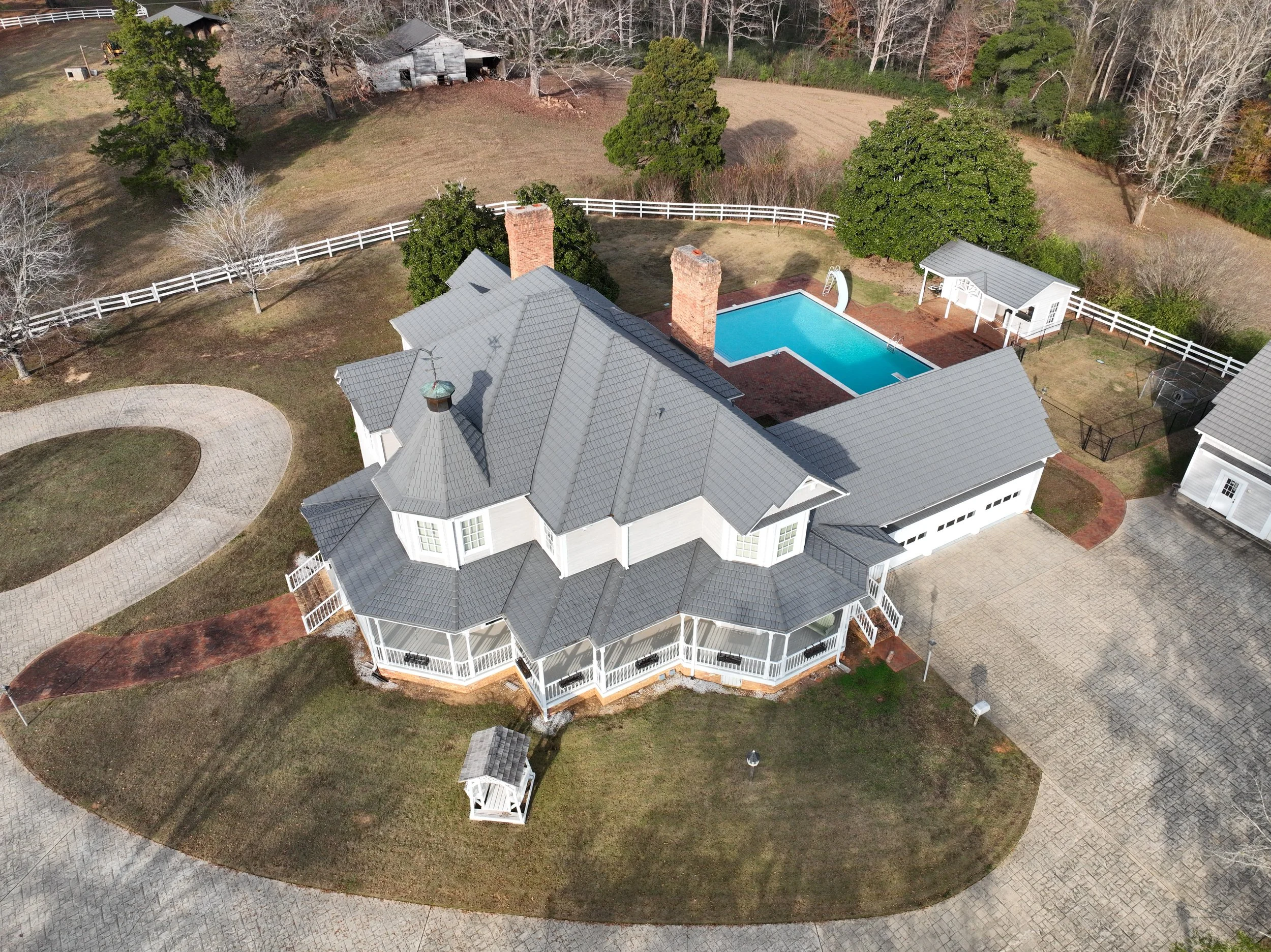 Aerial view of a large white house with gray roof, surrounded by a driveway, lawn, and trees. In the backyard, there is a swimming pool with a slide, a small shed, and a gazebo, all enclosed by a white fence.