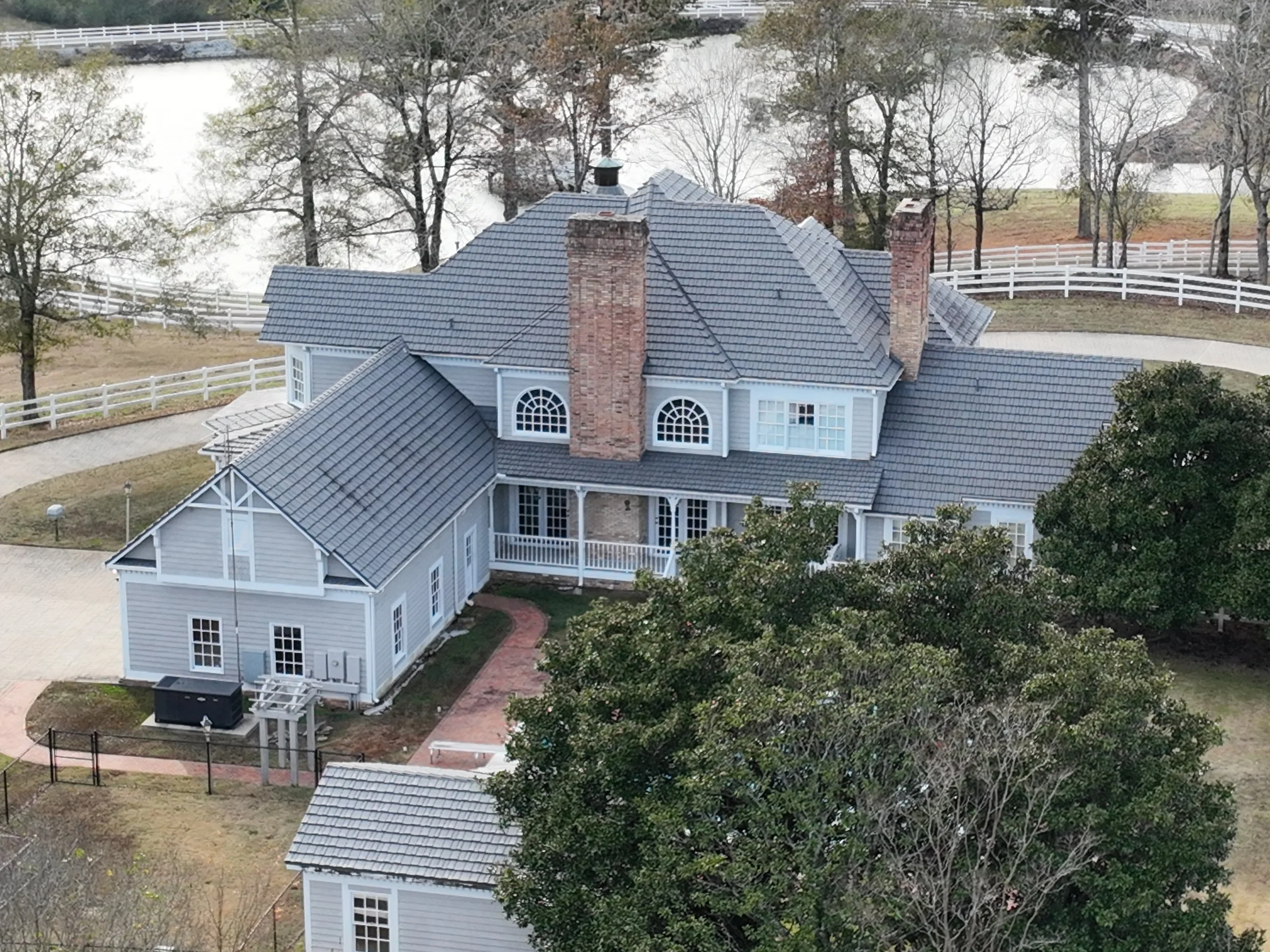 Aerial view of a large traditional house with a gray tiled roof, brick chimneys, surrounded by trees and a white fence, near a body of water.