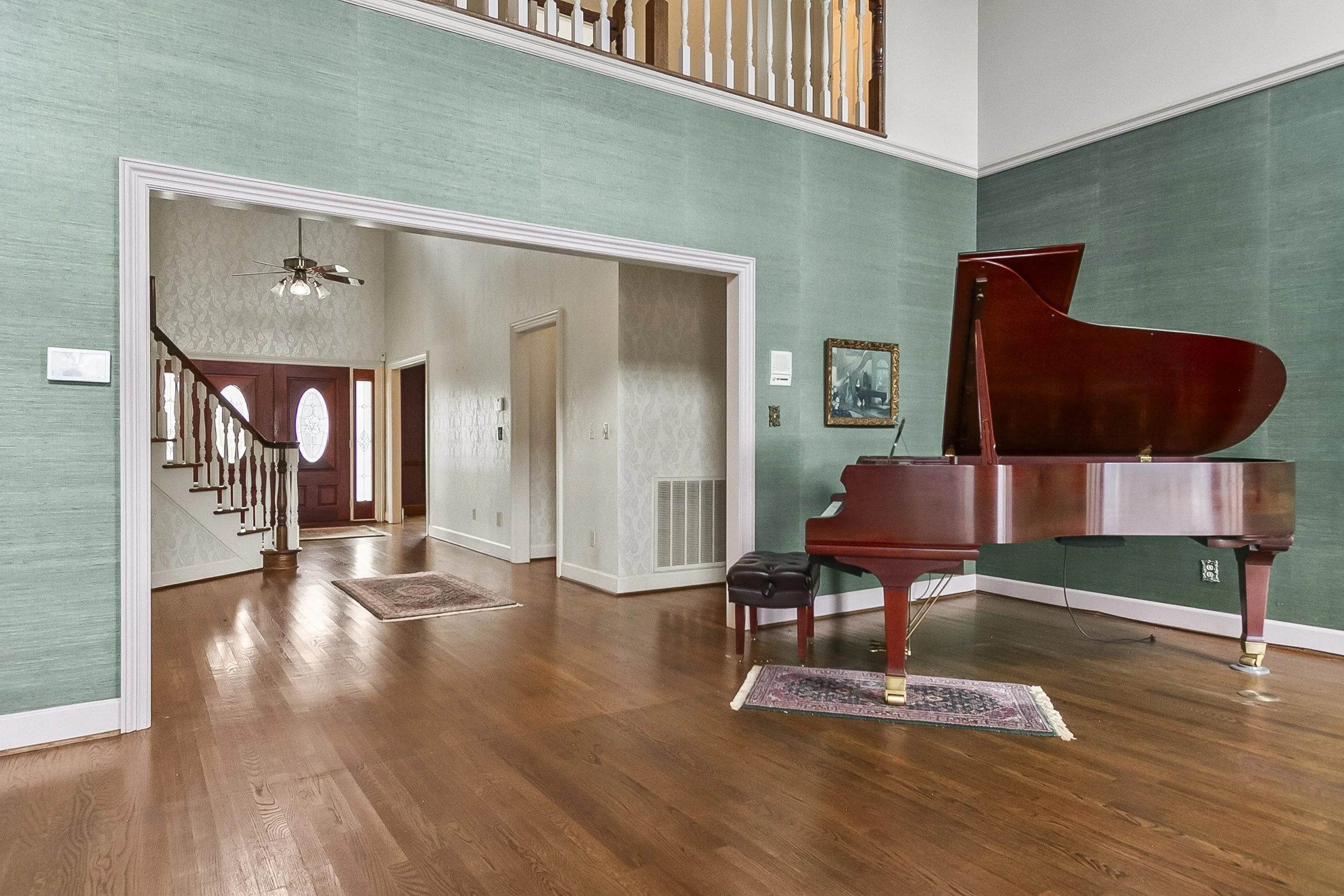A living room featuring a large wooden grand piano against a green textured wall, with a small rug underneath it. The room has hardwood flooring and a doorway leading into an entryway with a front door visible in the background. There is a staircase 