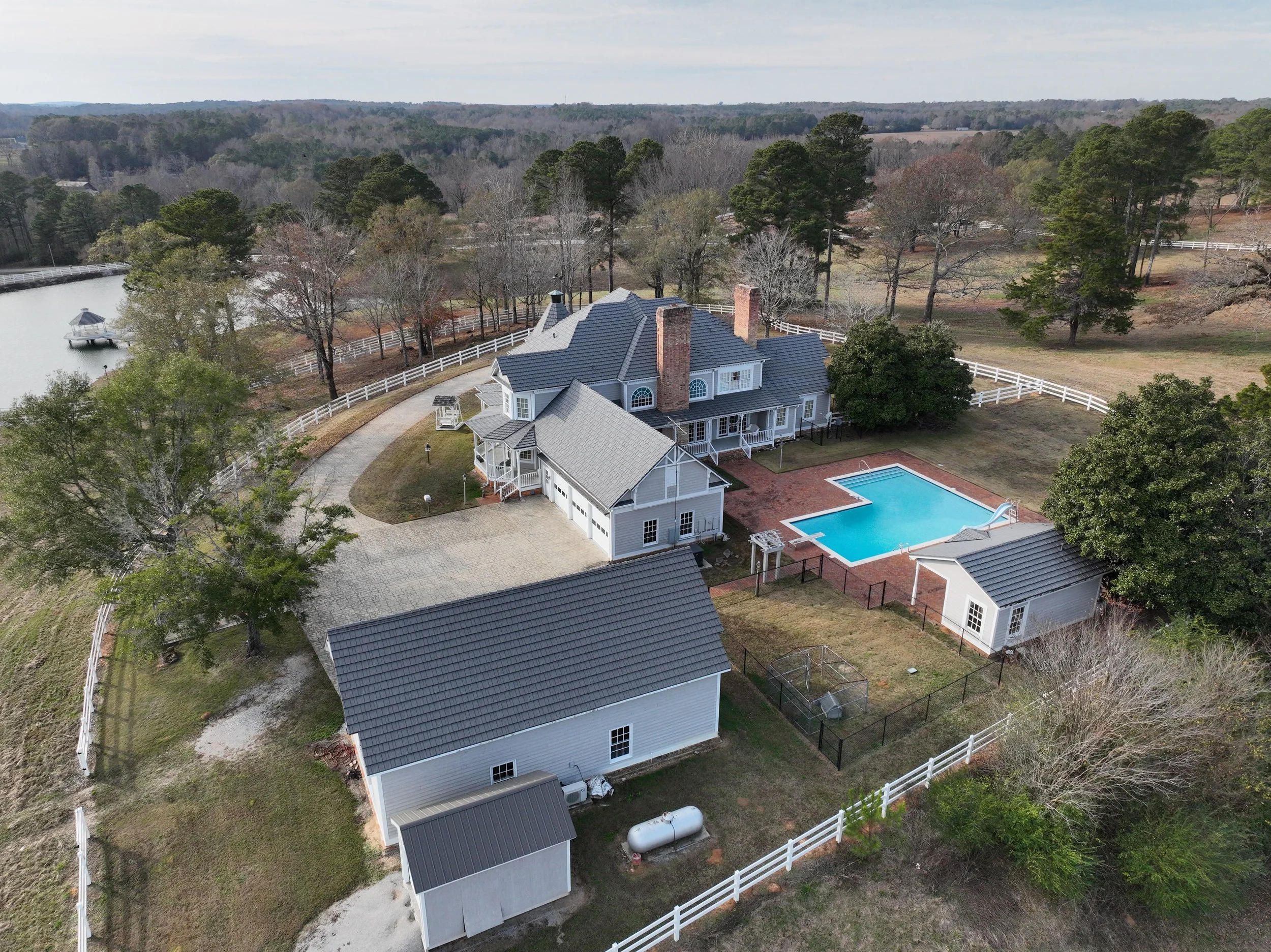 Aerial view of a large house with grey roof, swimming pool, and backyard, surrounded by trees and fenced yard, beside a lake with a dock and boat, in a rural setting.