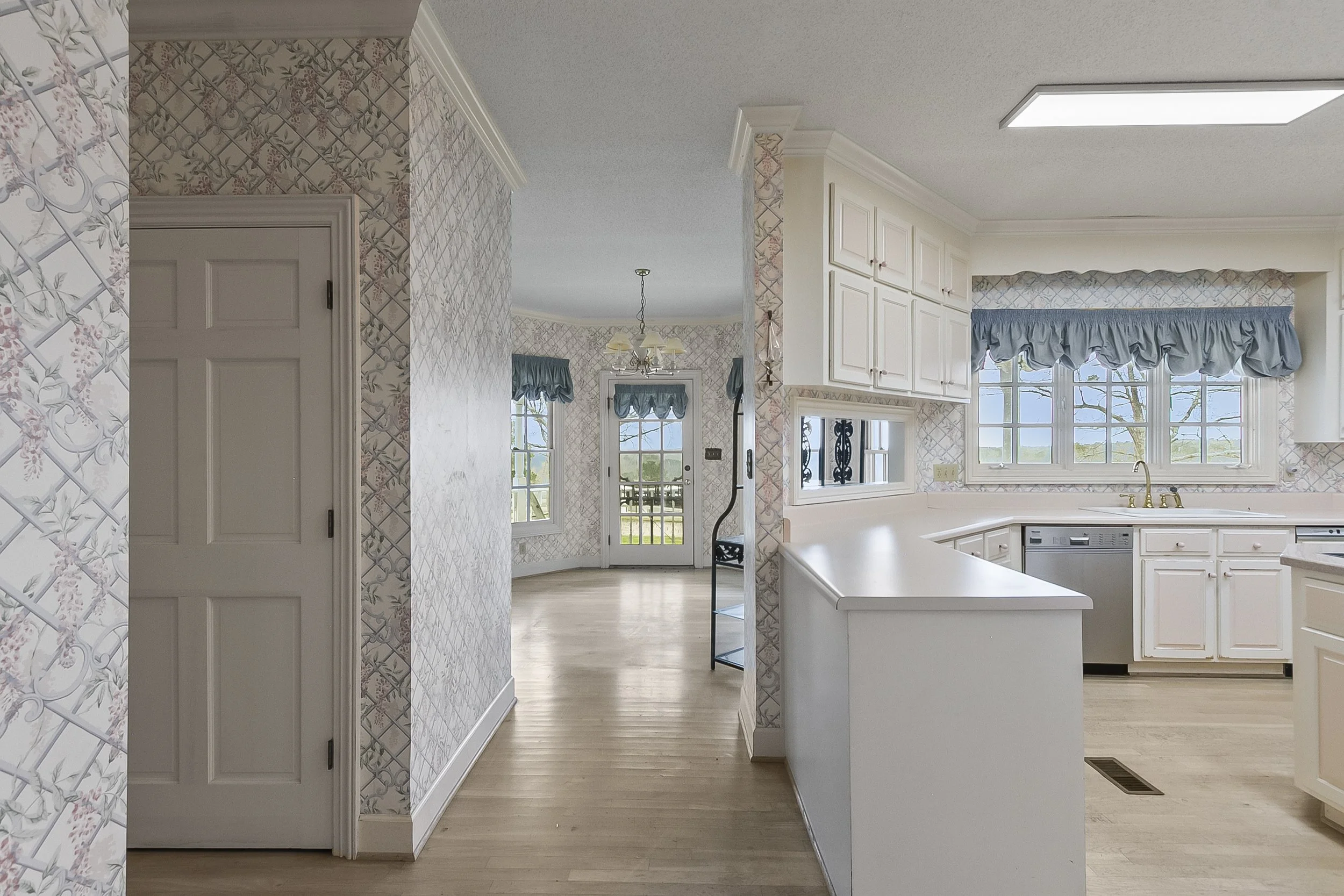 Interior of a bright, spacious kitchen with white cabinetry, a window with a valance, and a view to a dining area and outside patio.