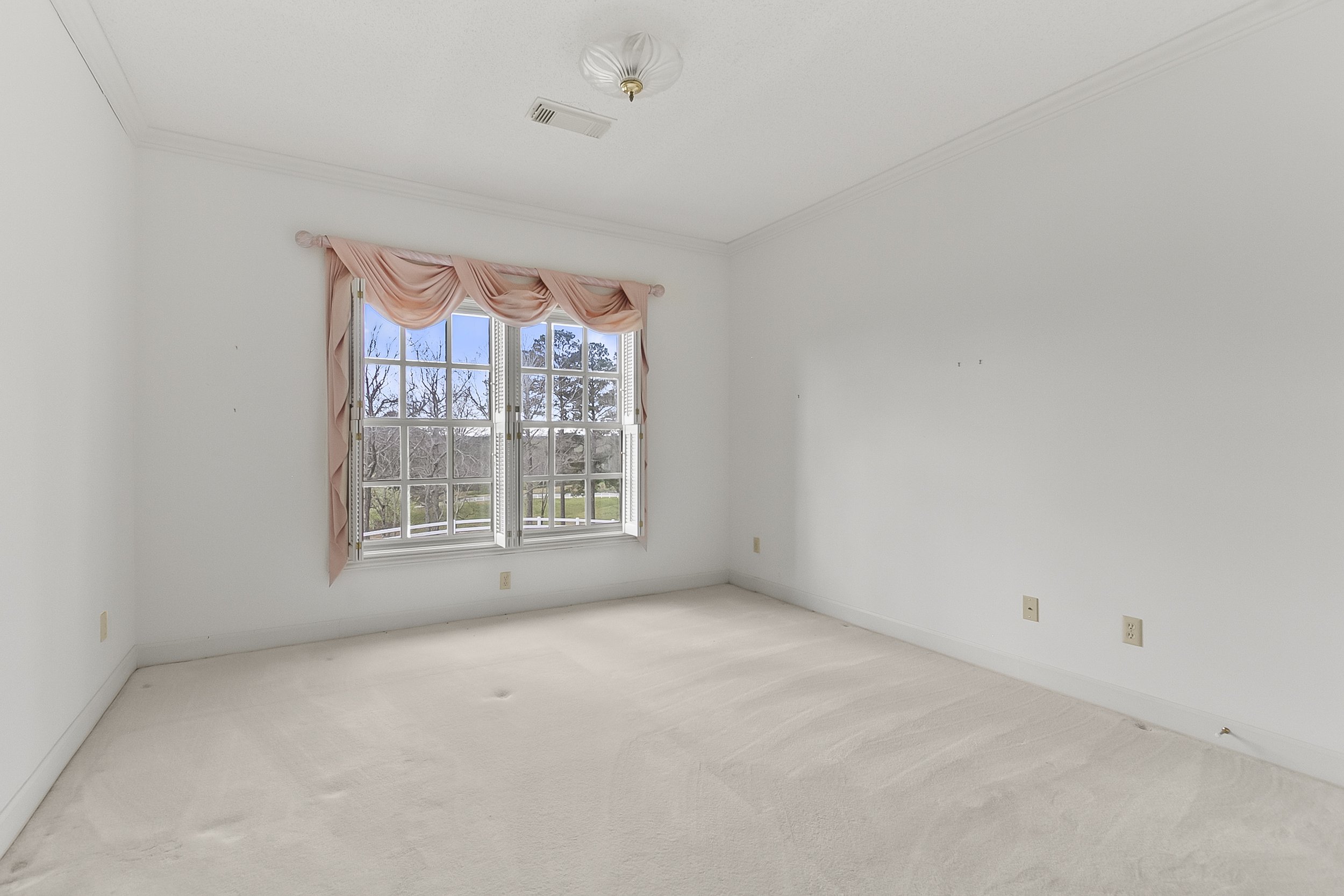Empty room with white walls, a large window with pink curtains, carpeted floor, ceiling light fixture, and view of trees outside.
