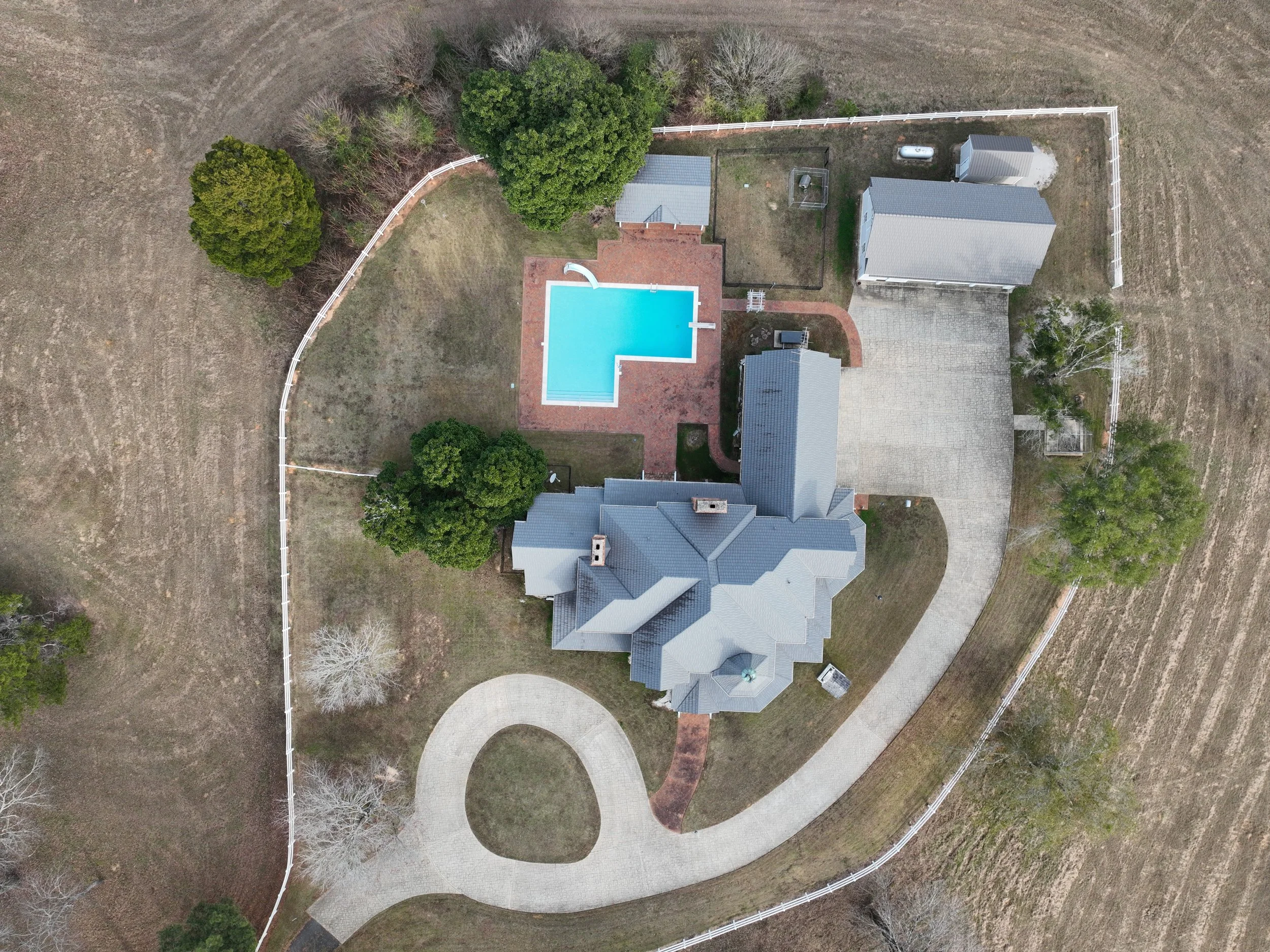 Aerial view of a large house with a gray roof, a swimming pool with a red brick patio, surrounded by trees, grass, and a fenced yard, with a curved driveway.