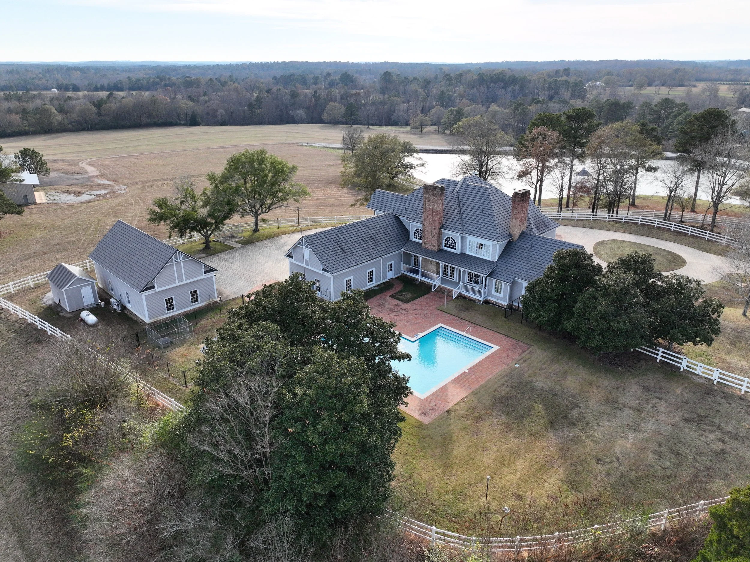 A large two-story house with a swimming pool in the backyard, surrounded by trees and white fences, with a pond and fields in the background.