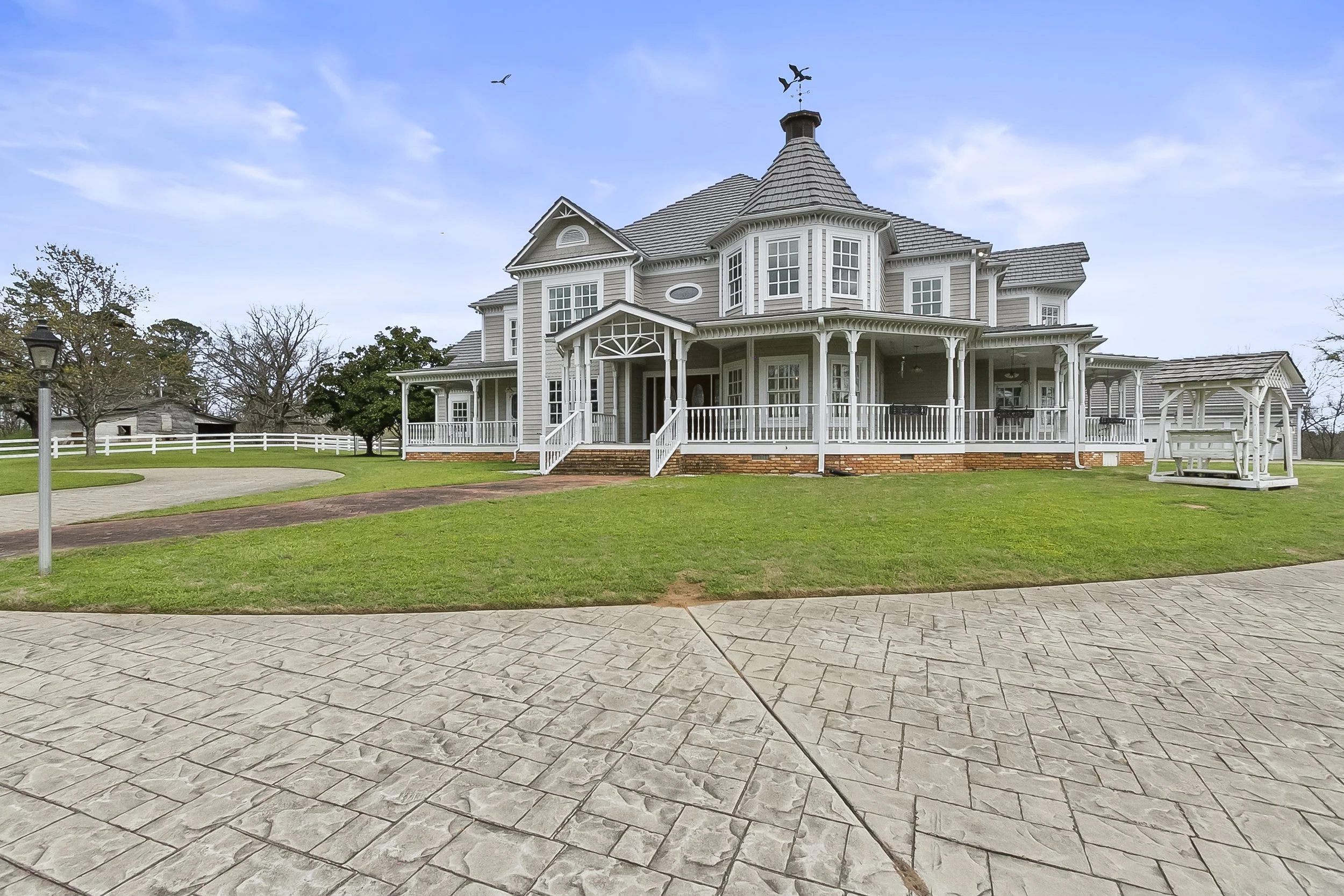 Large Victorian-style house with a wraparound porch, surrounded by a well-maintained lawn and paved driveway, under a partly cloudy sky.