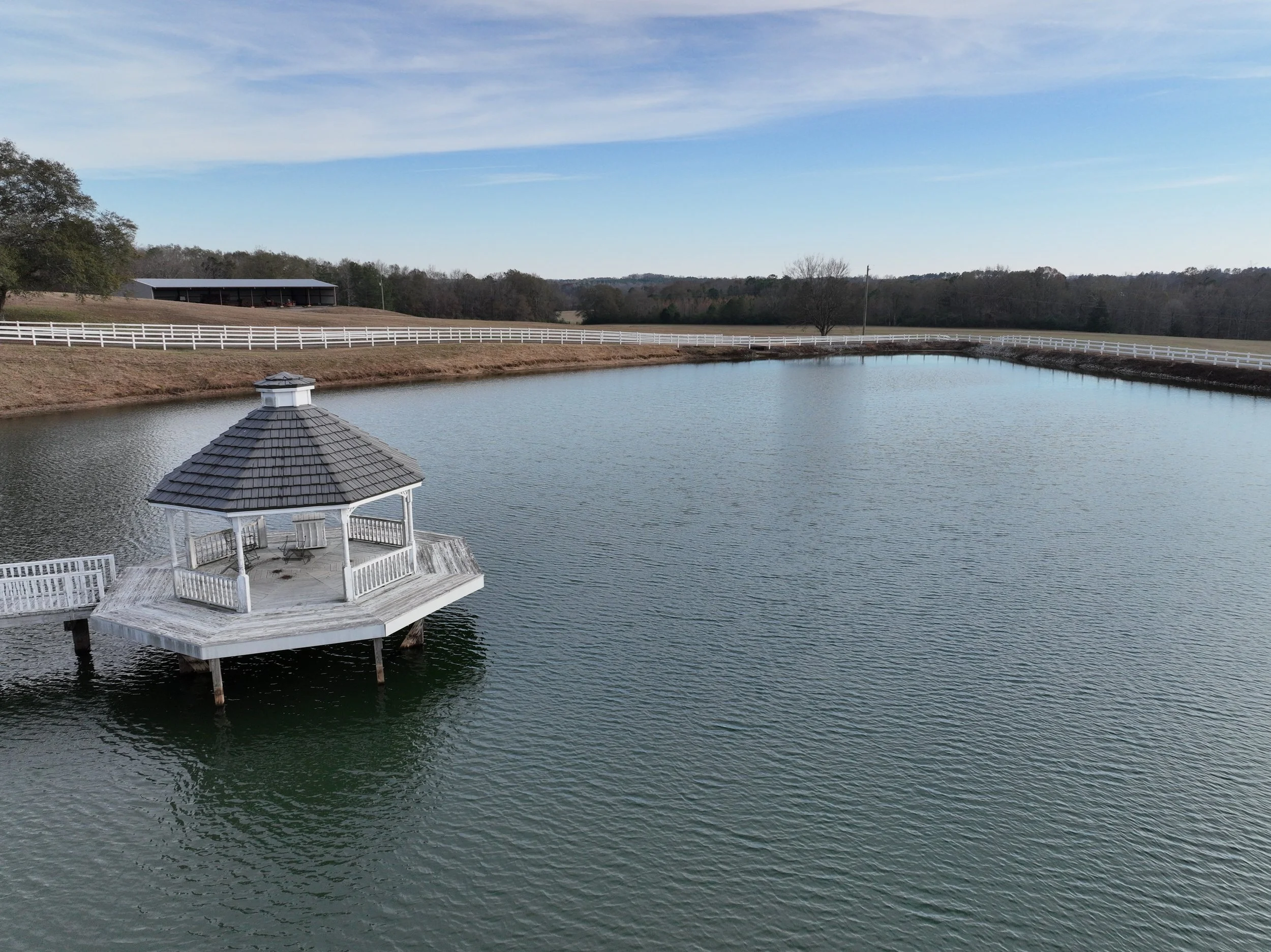 A peaceful pond with a wooden gazebo extending over the water, surrounded by a white fence, with a large barn and trees in the background under a partly cloudy sky.