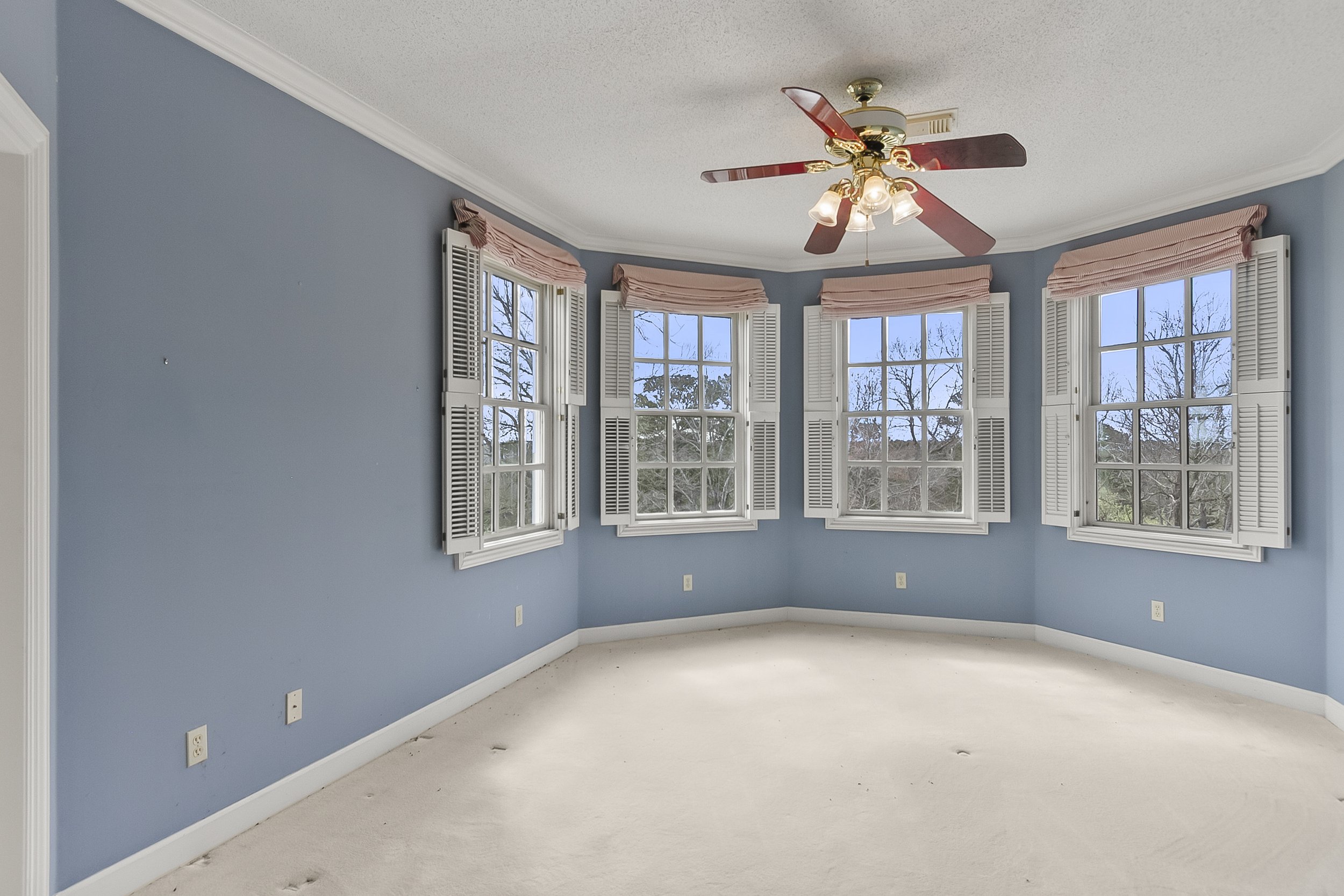 Empty blue-walled room with four large windows with white shutters and pink valances, a ceiling fan with five blades and five light bulbs, and a beige carpet.