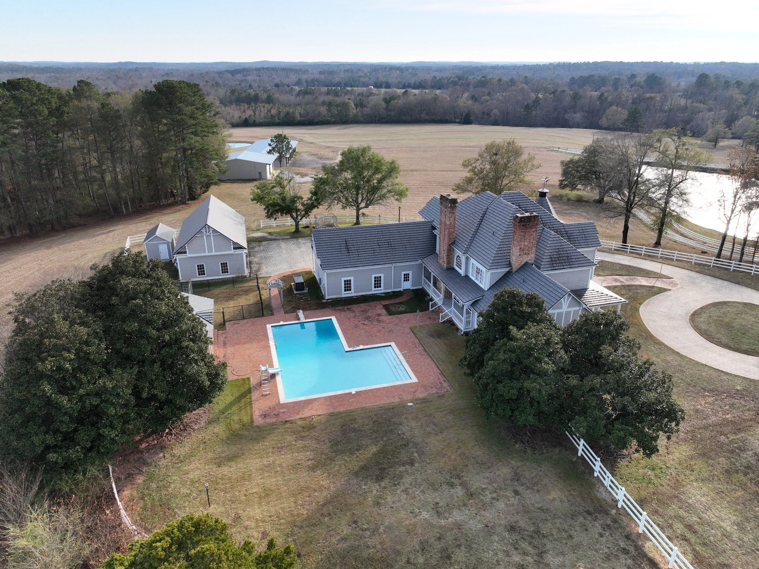 Aerial view of a large house with a swimming pool in the backyard, surrounded by trees, outbuildings, and a rural landscape with fields and trees.