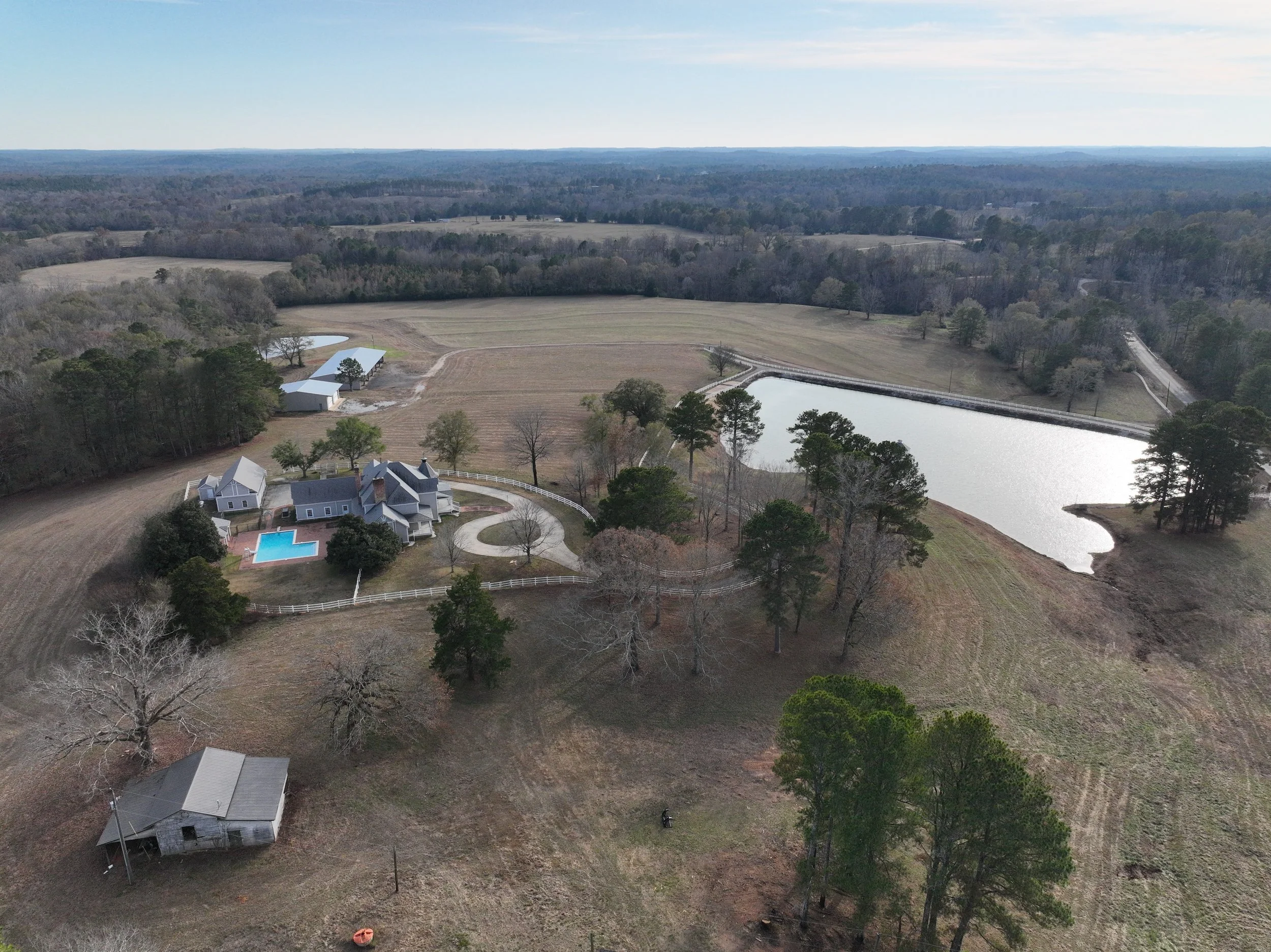Aerial view of a rural property with a large house, swimming pool, pond, and open fields surrounded by trees, with farmland and wooded areas extending to the horizon.
