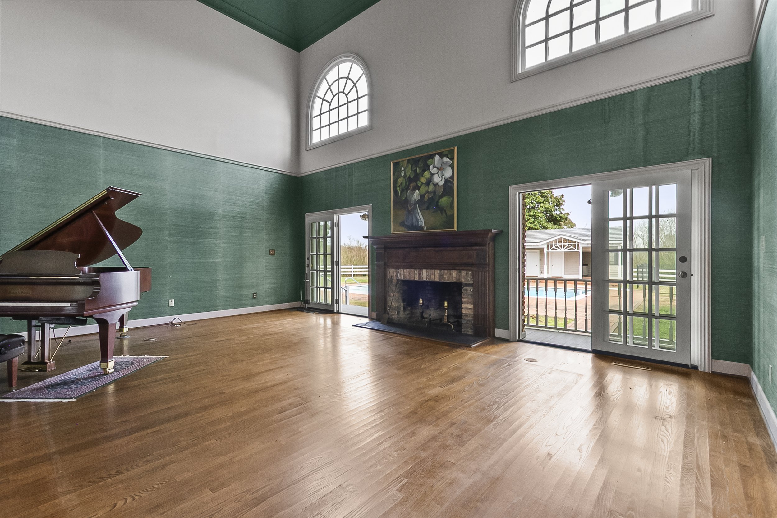 Living room with green walls, wooden floor, a piano on the left, a fireplace with a painting above it in the center, and two glass doors leading outside, one on each side of the fireplace.