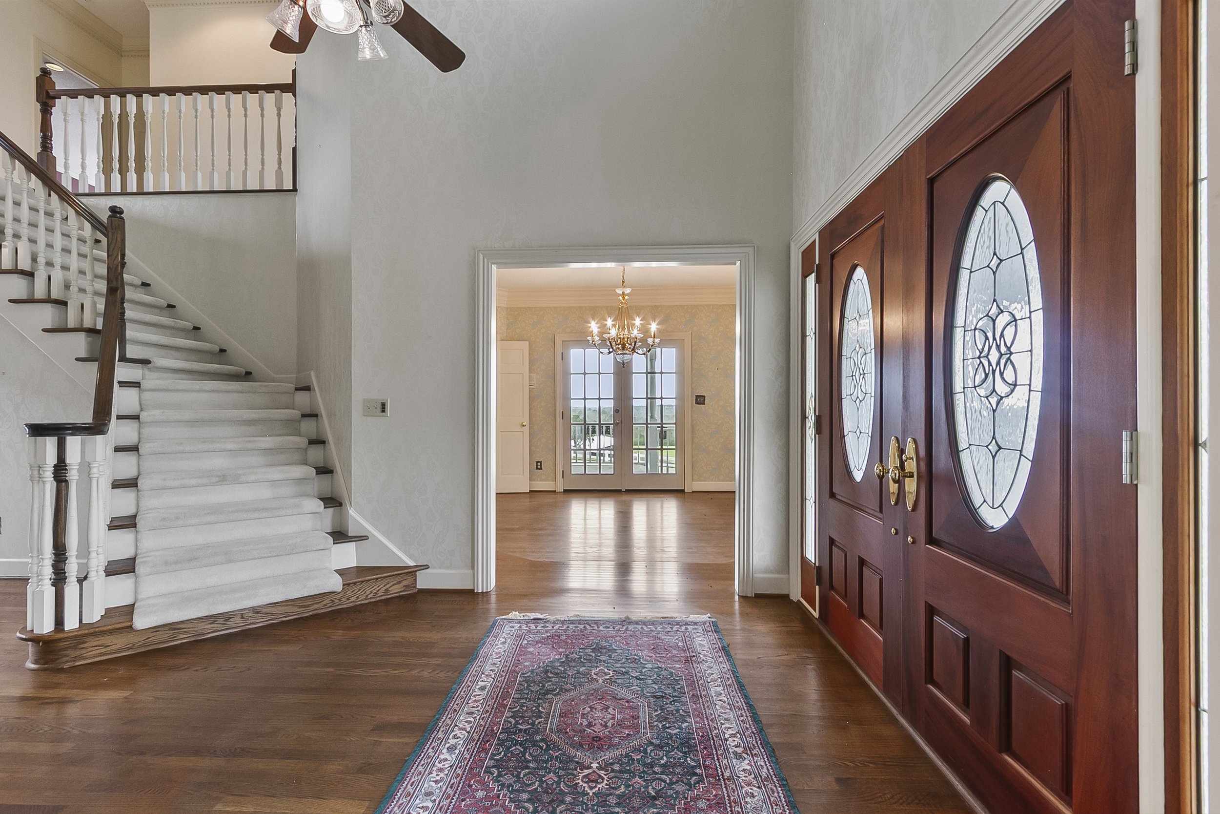 Entrance of a home with wooden double doors with oval glass panels, a patterned rug on hardwood flooring, and a view into a dining room with a chandelier, chandelier, and French doors leading outside.
