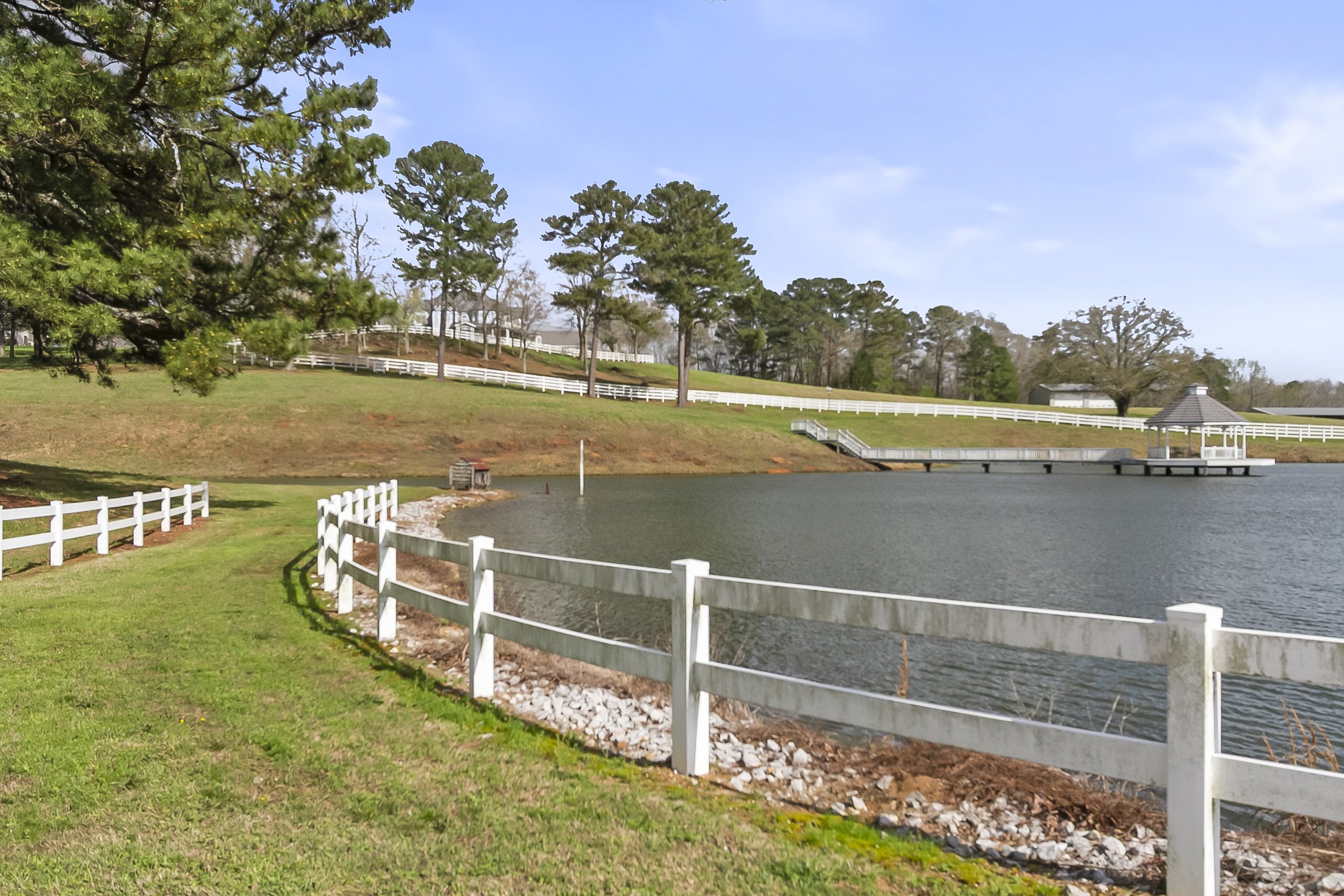 A peaceful lakeside scene with a white wooden fence along the grassy shore, trees in the background, a small dock with a pavilion over the water, and a blue sky with some clouds.