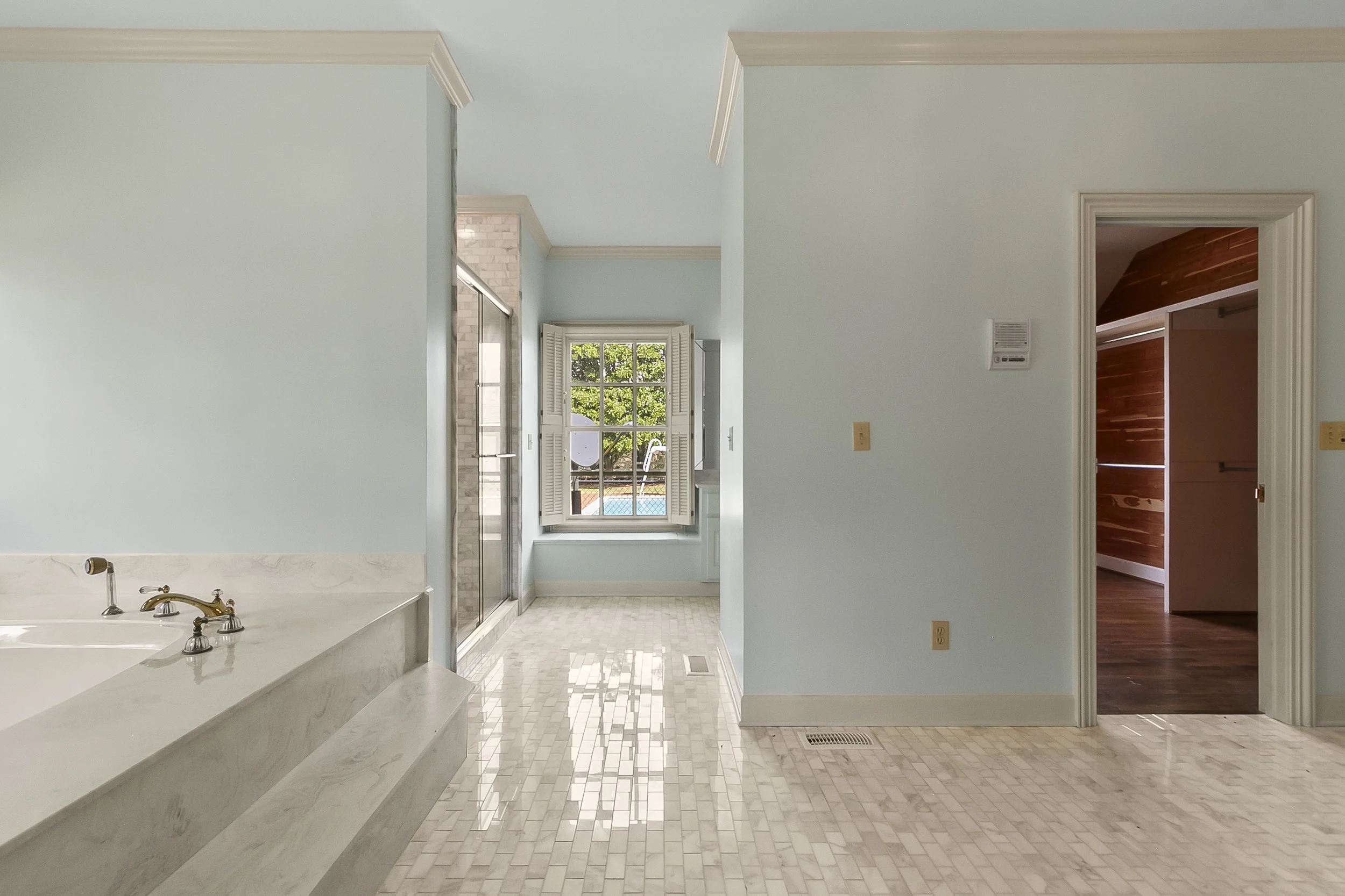 Empty bathroom with light blue walls, tile flooring, a large window with open shutters, a glass shower stall, a bathtub with a marble surround, and a doorway leading to a room with wooden floors and wood-paneled walls.