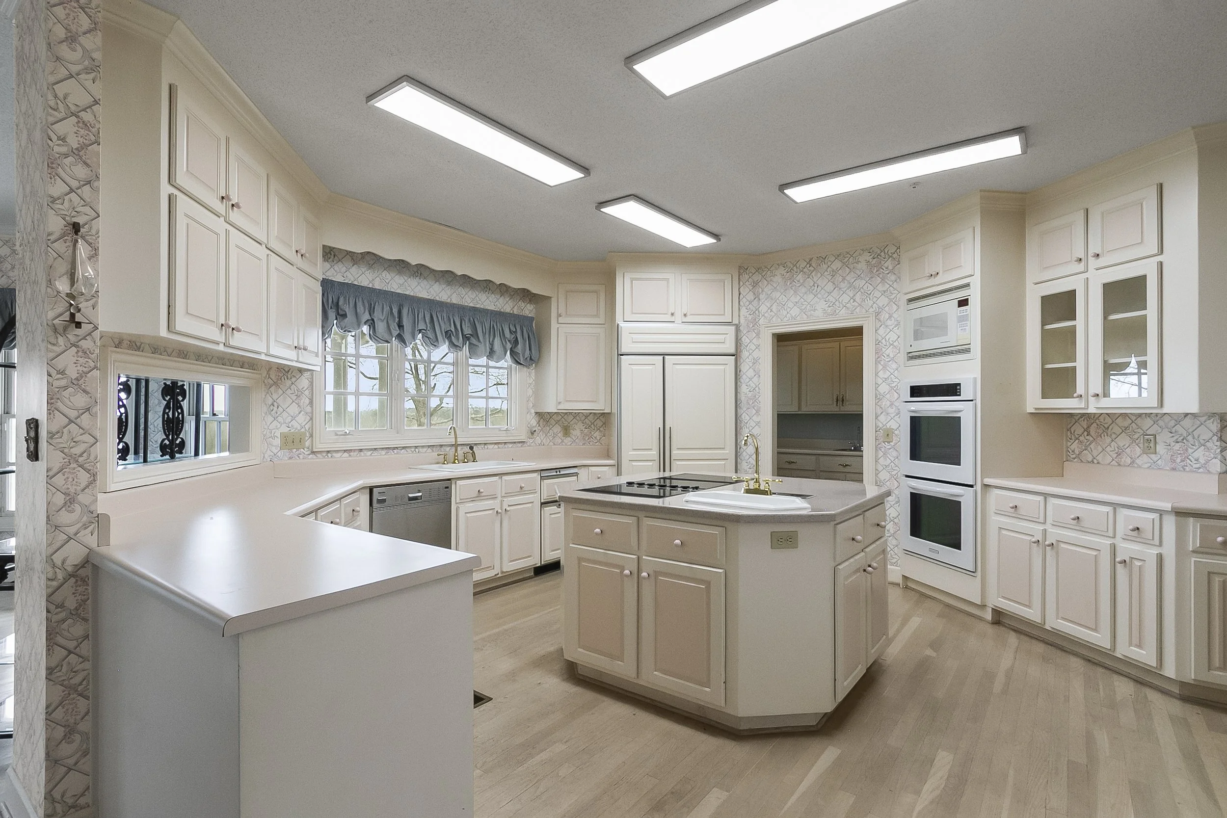 A spacious, bright kitchen with white cabinetry, a central island with a cooktop, and multiple windows with blue valances, featuring a light wood floor and built-in appliances.