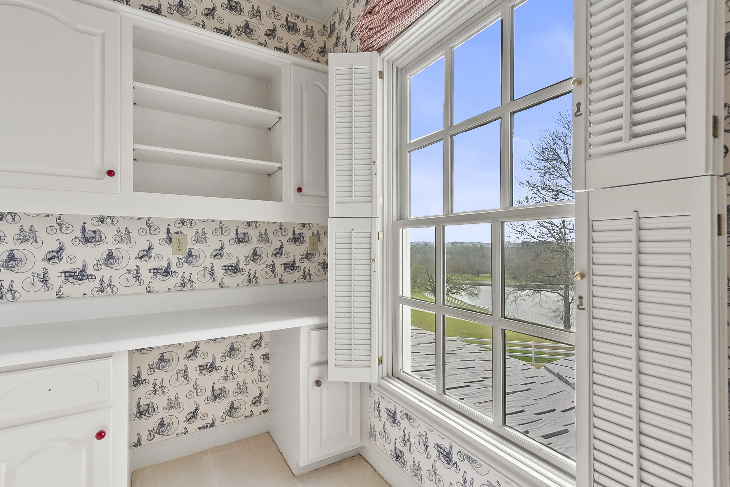 White built-in cabinets with open shelves and closed doors, decorated with bicycle-themed wallpaper, next to a large window with shutters overlooking a scenic outdoor landscape with a lake and trees.