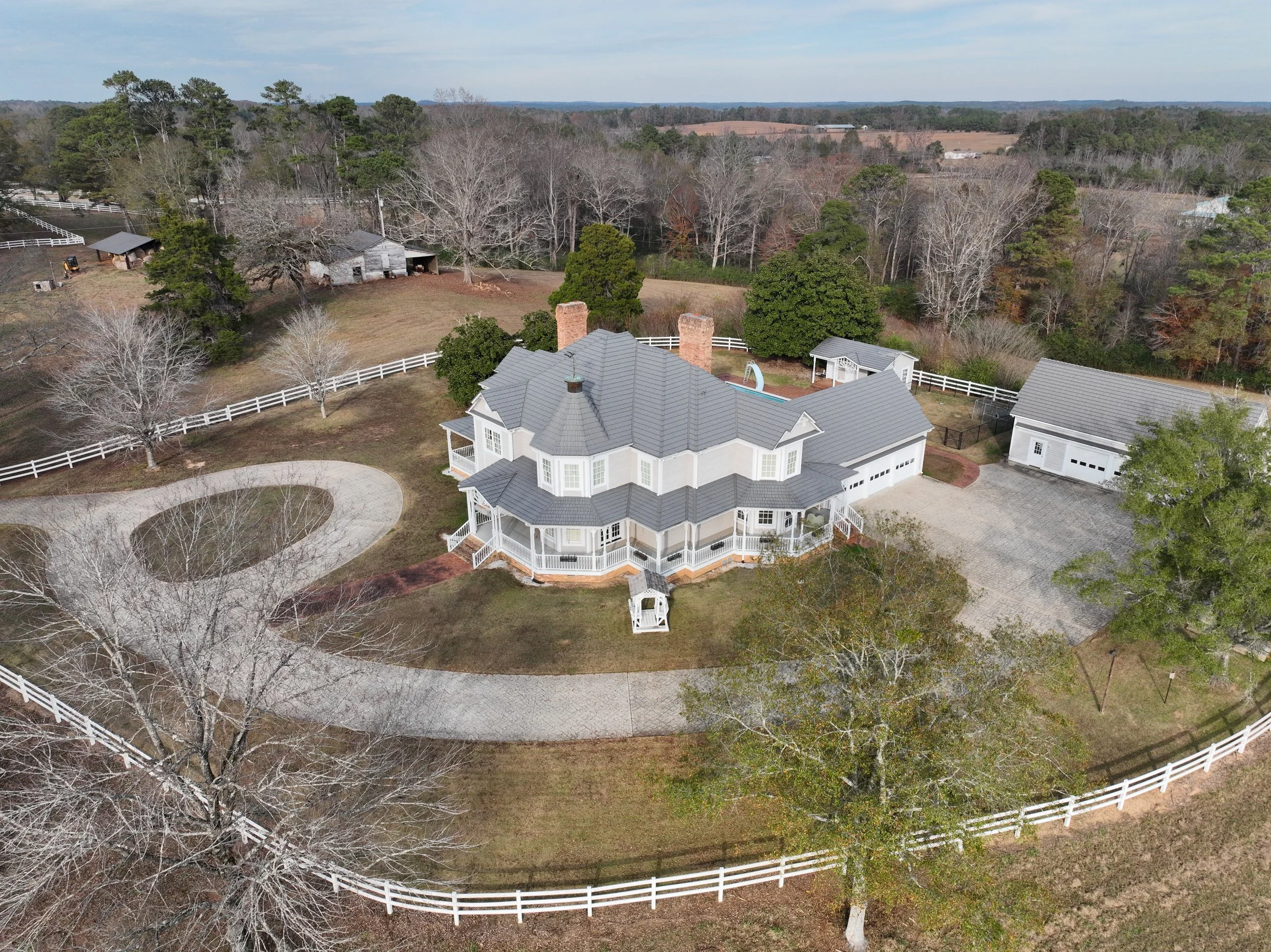 A large white house with a gray roof, a circular driveway, surrounded by trees and a white fence, on a rural property.
