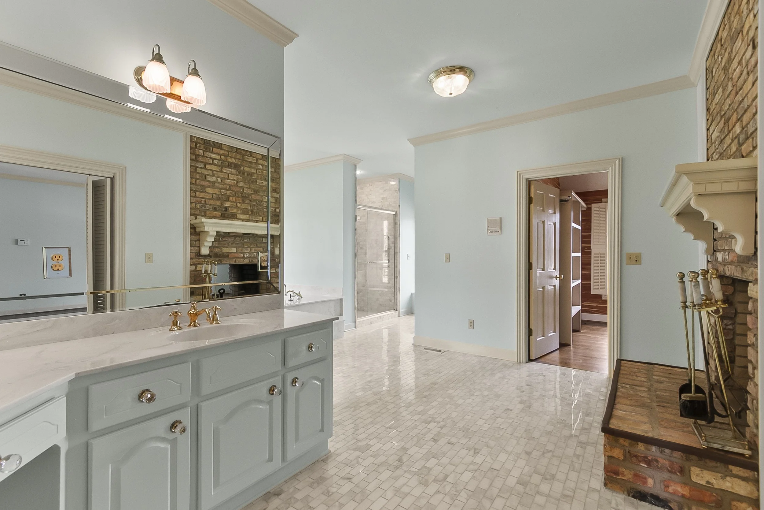 Living space with light blue walls, a brick fireplace, and a bathroom with a shower. There are doors leading to other rooms, and a mirrored cabinet above a light gray vanity with gold fixtures.
