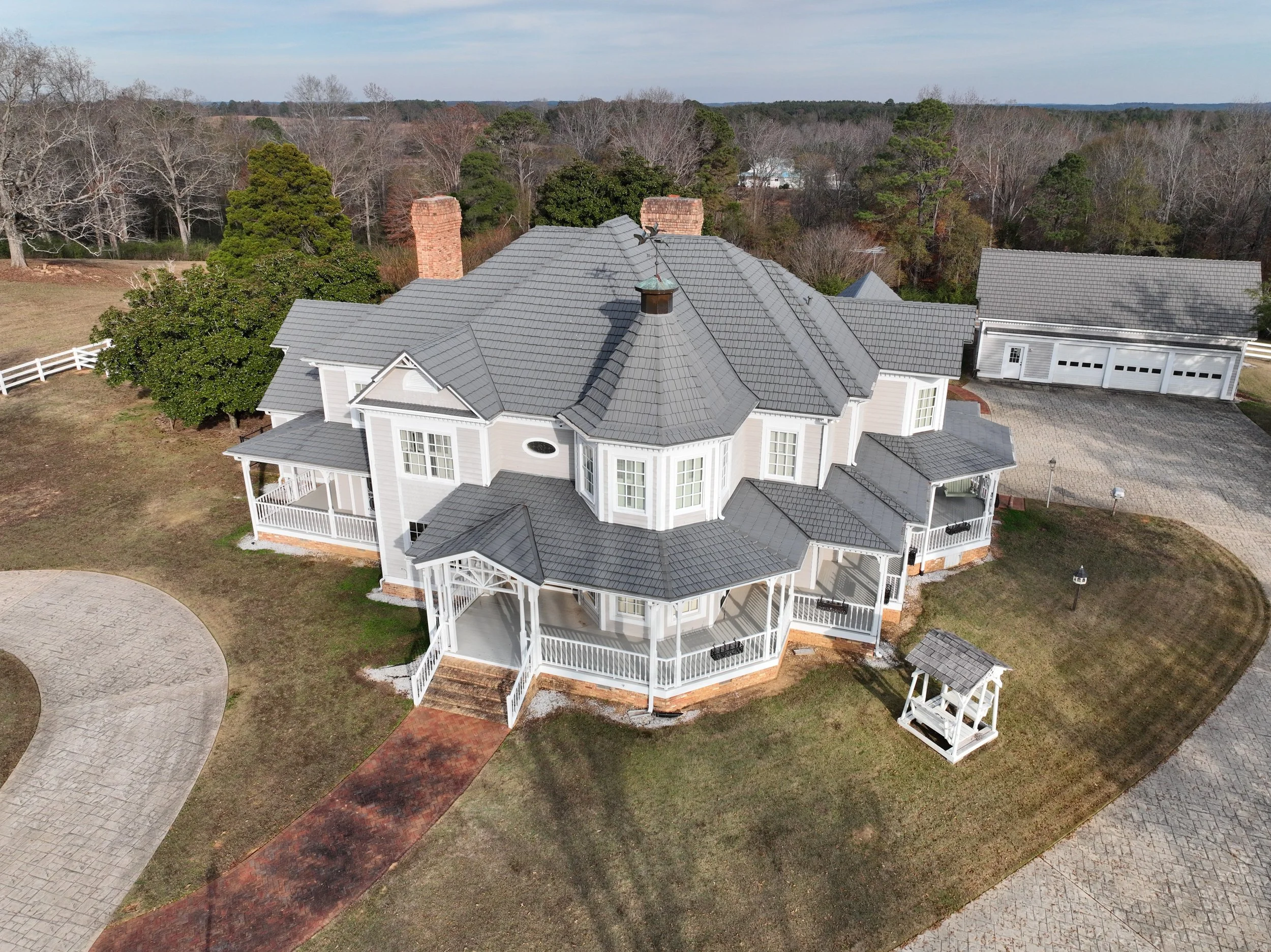 An aerial view of a large, white, Victorian-style house with a gray tiled roof, multiple dormer windows, and a wraparound porch, surrounded by trees and a driveway.
