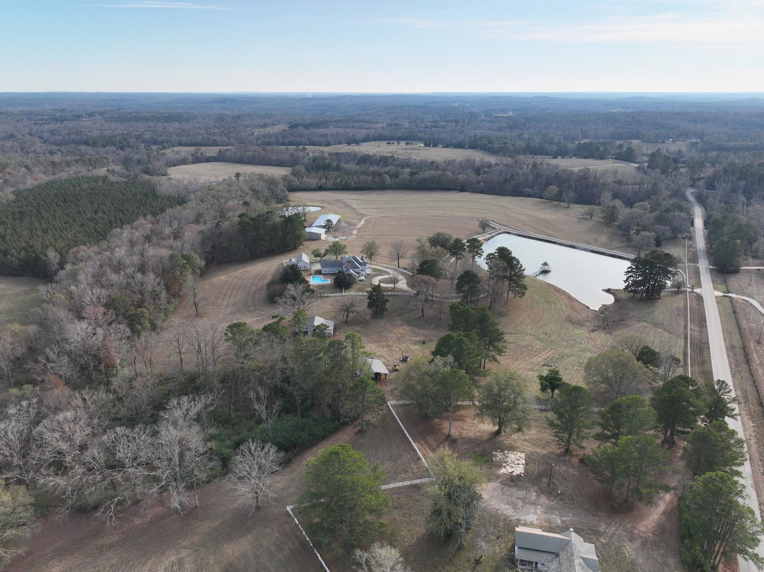 Aerial view of a rural property with a house, swimming pool, pond, trees, fields, and a road.
