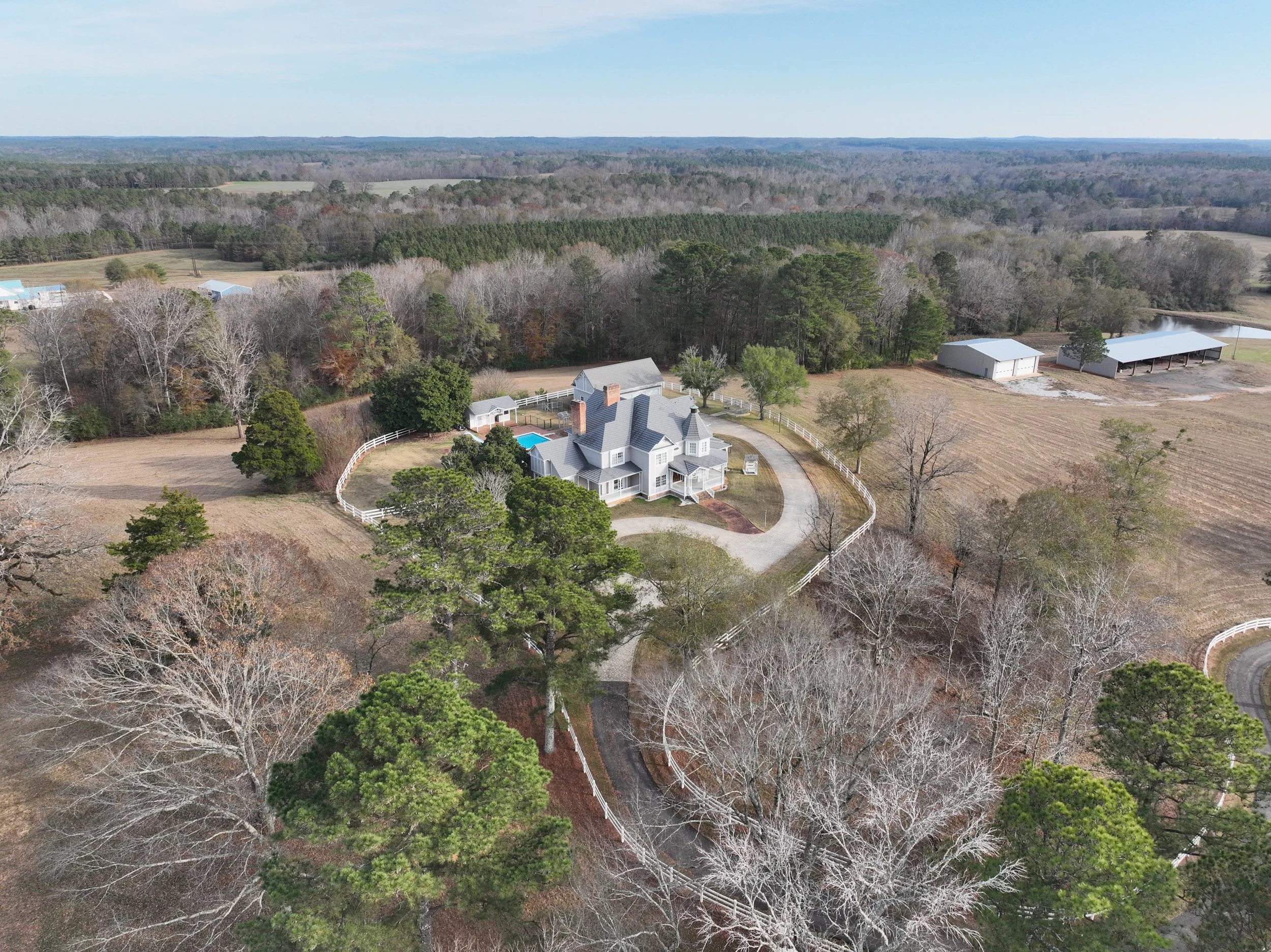 Aerial view of a large house with a driveway, swimming pool, and surrounding trees, located in a rural area with fields and additional buildings nearby.