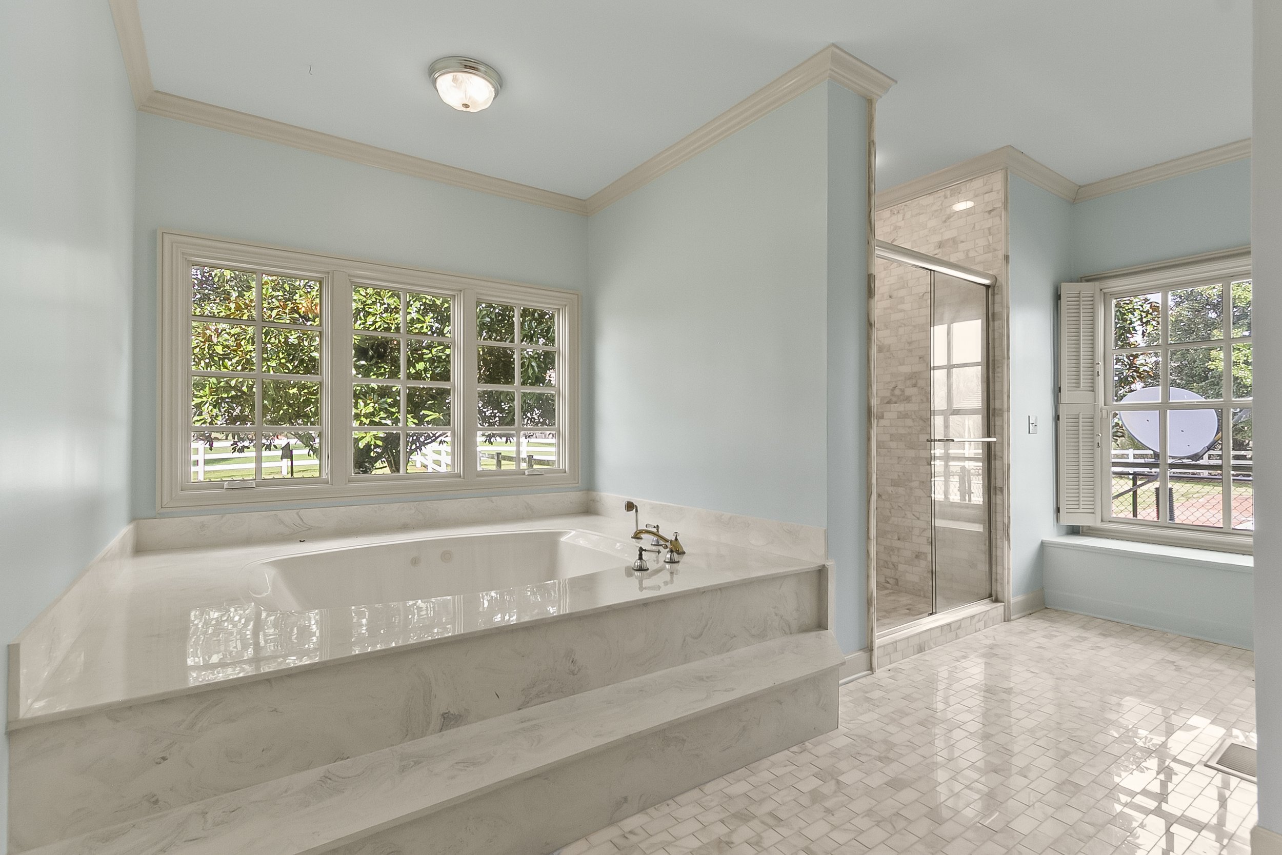 Bathroom with a large bathtub, marble surround, and a window showing greenery outside. There is a glass-enclosed shower with marble walls and a window with shutters. The floor is tiled with small white tiles, and the walls are painted light blue.