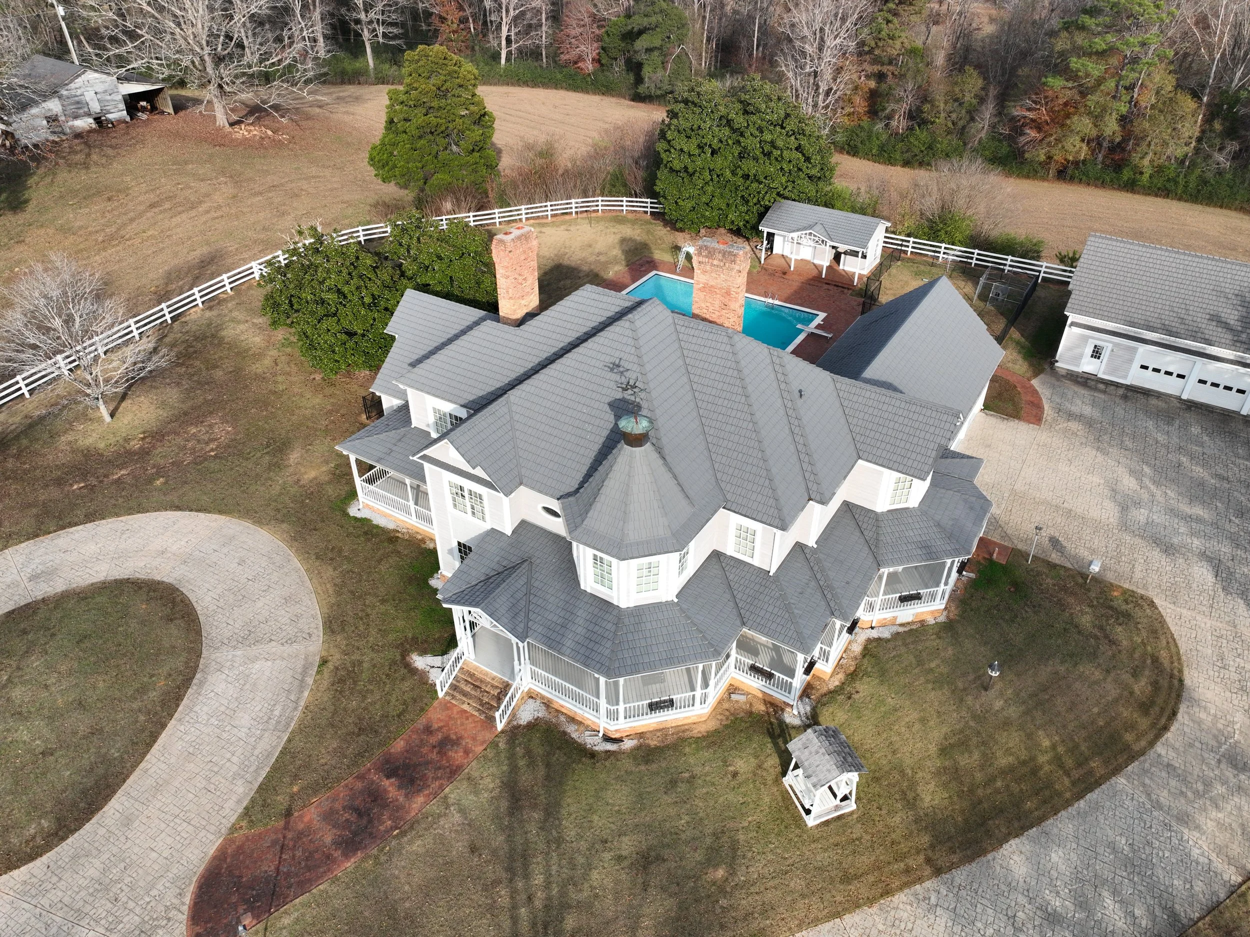 Aerial view of a large white house with gray roof, surrounding yard with curved driveway, swimming pool, and nearby shed, set in a spacious backyard with trees and fenced area.