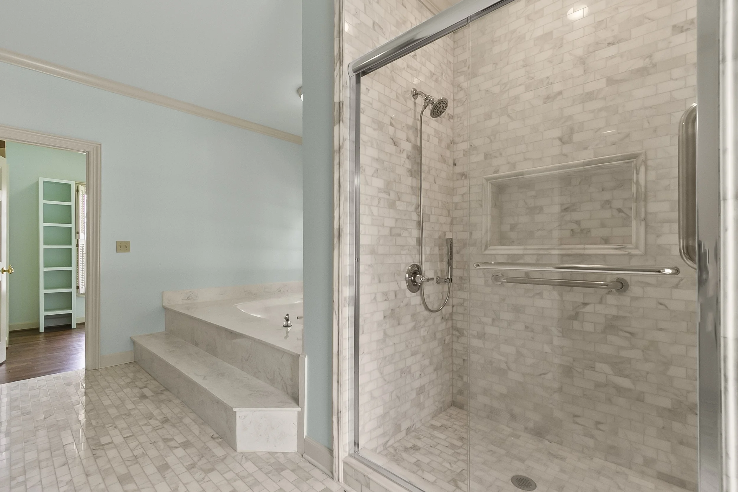 Bathroom with a walk-in shower with marble tiles and a built-in shelf, and a corner bathtub, with a light blue wall and doorway leading to another room.