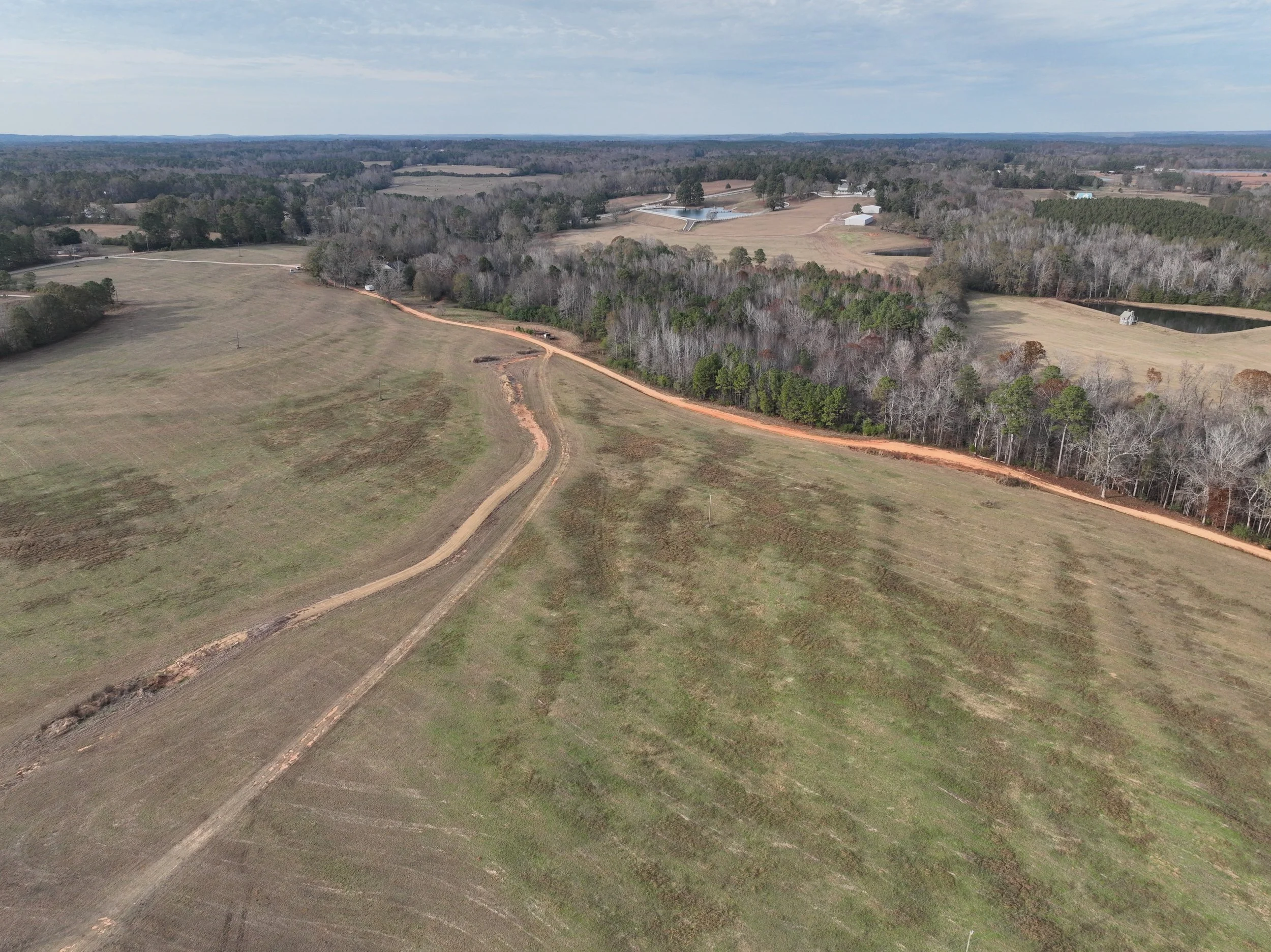 Aerial view of an open field with a dirt road winding through it, bordered by trees, and a pond in the background under a cloudy sky.
