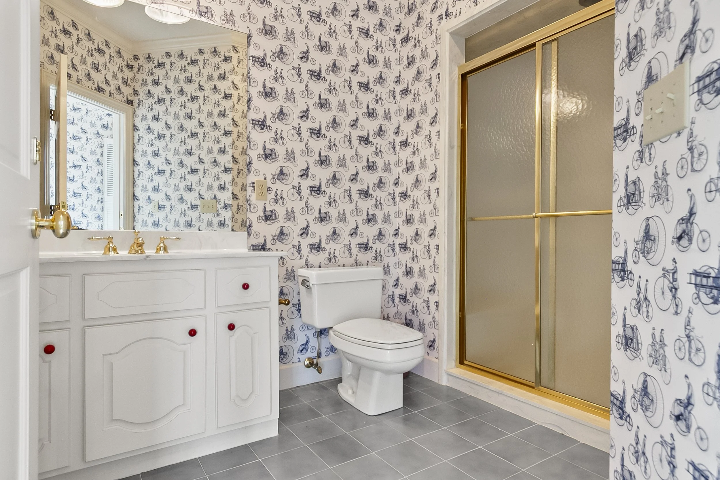 A bathroom with wallpaper featuring bicycles, a white vanity with a mirror and gold fixtures, a toilet, and a glass shower with gold trim.