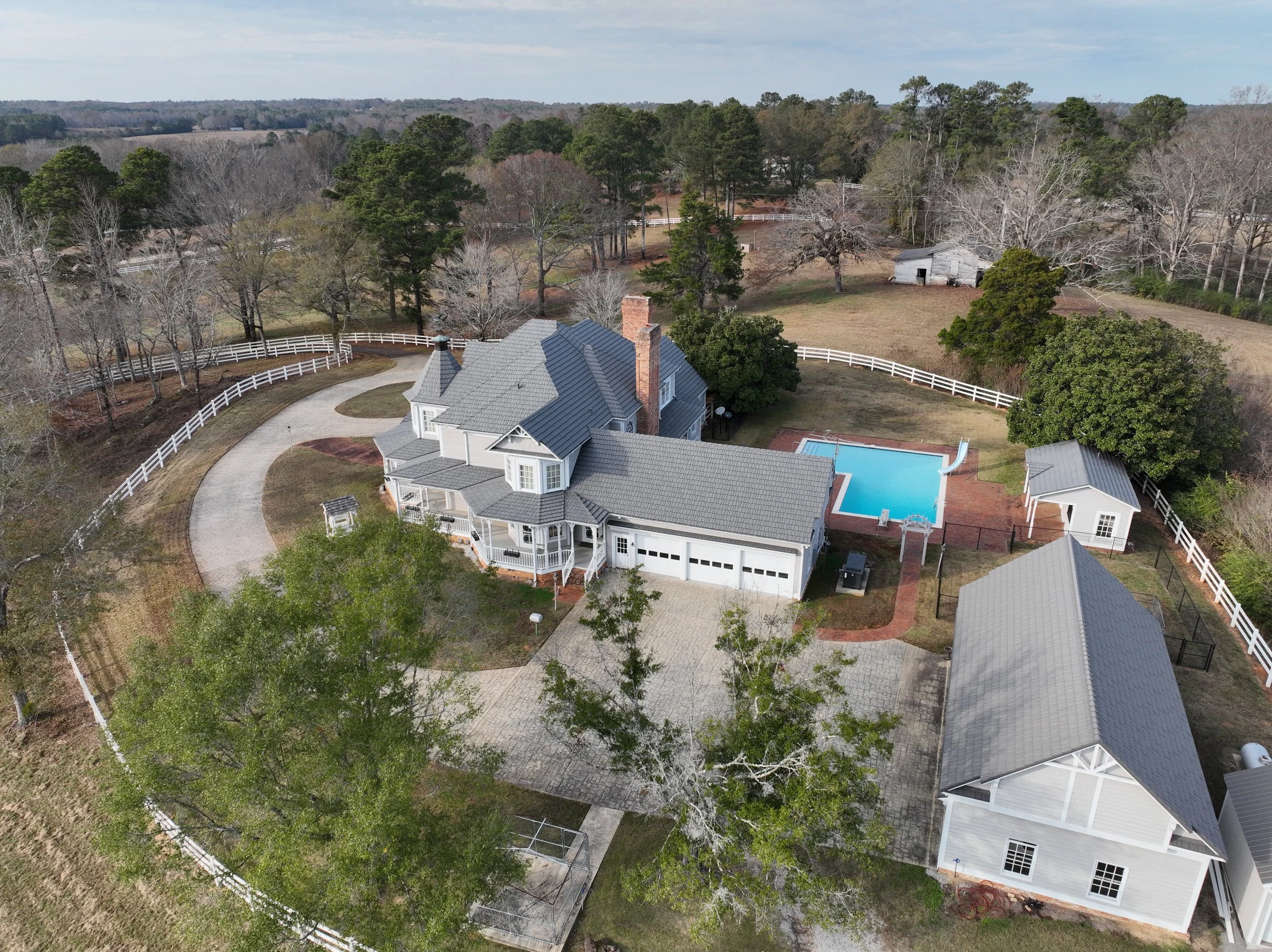 A large white house with a gray roof, a chimney, a driveway, a swimming pool, and a lawn, surrounded by trees and a white fence.
