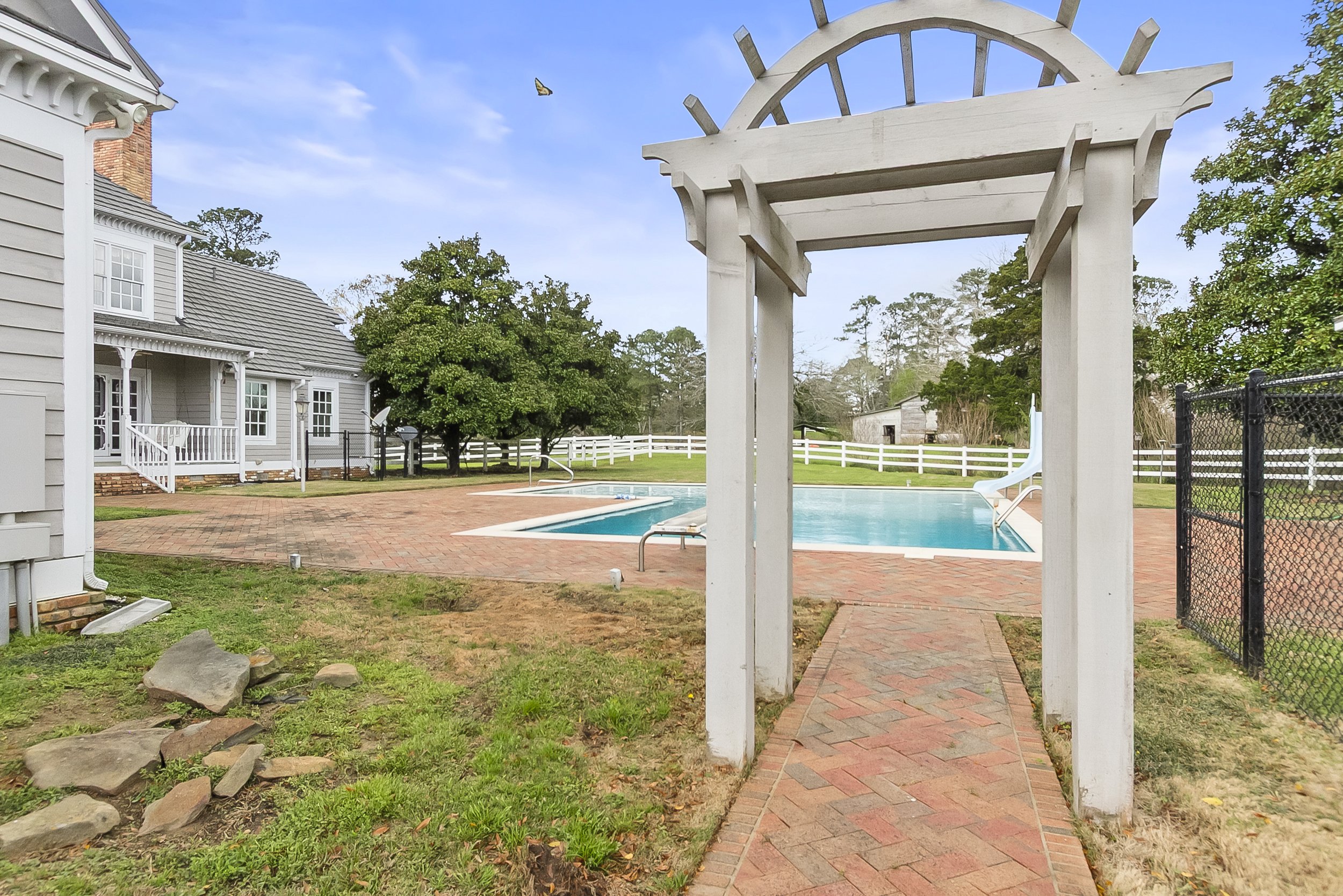 View of a backyard with a swimming pool, white fencing, a slide, and a house, with a wooden archway over a brick pathway leading to the pool