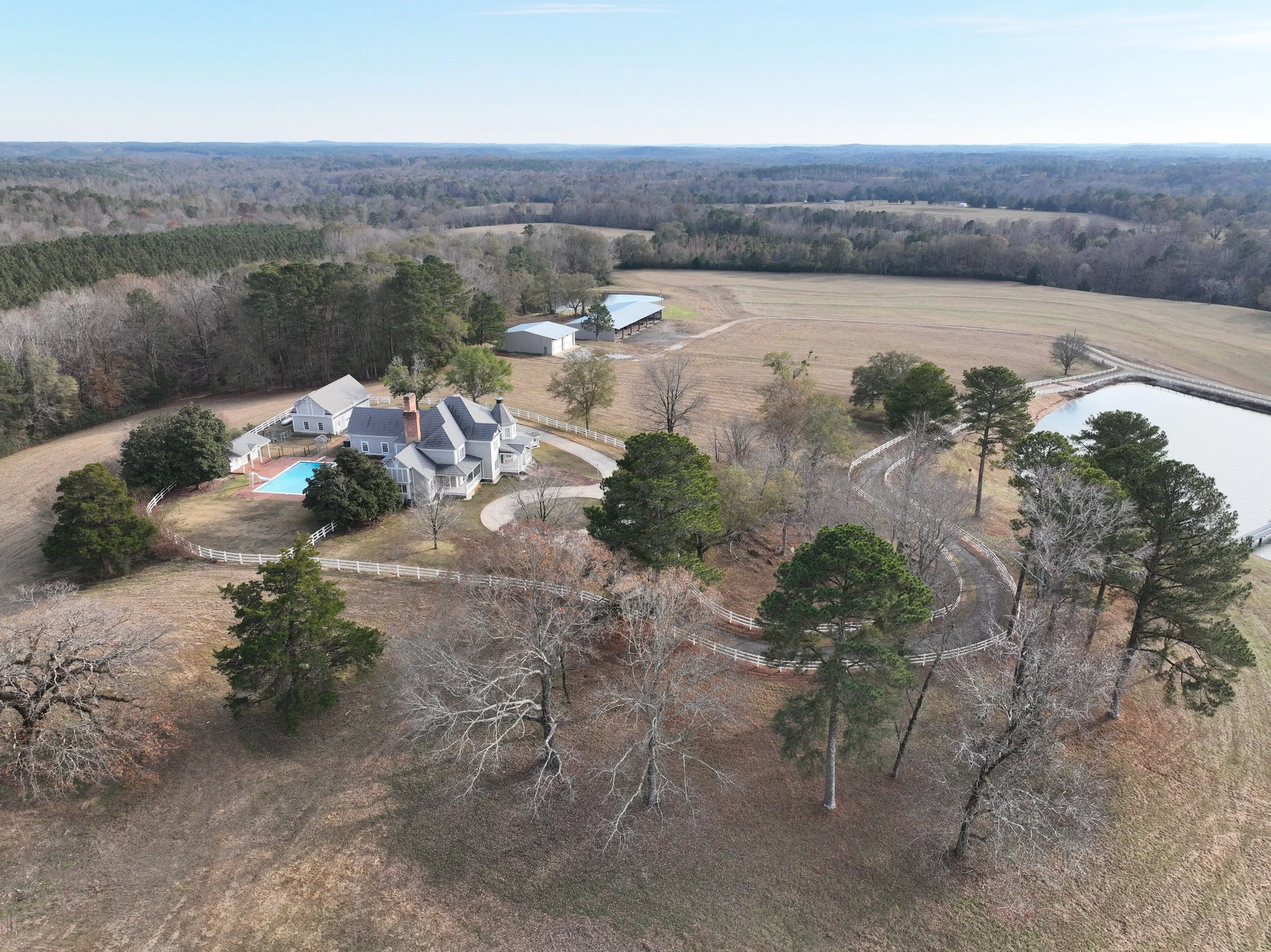 A large country estate featuring a white house with a gray roof, outdoor swimming pool, surrounded by a white fence, tall trees, open fields, a pond or lake, and distant wooded hills under a clear sky.