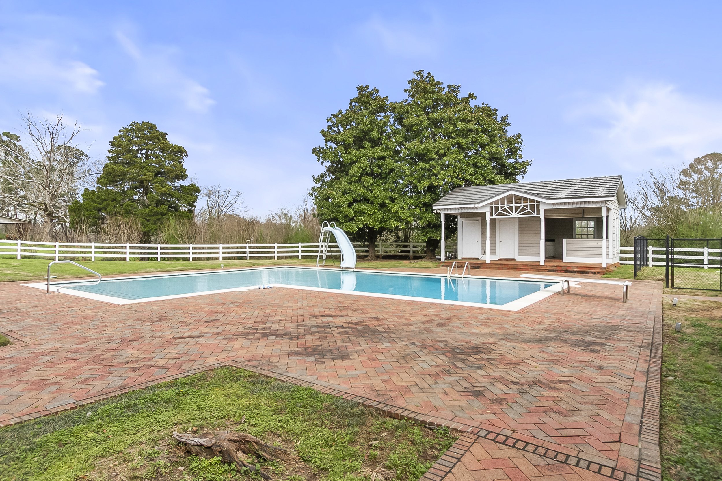 Residential backyard with a swimming pool, a small pool house, surrounding trees, a white fence, and a brick patio