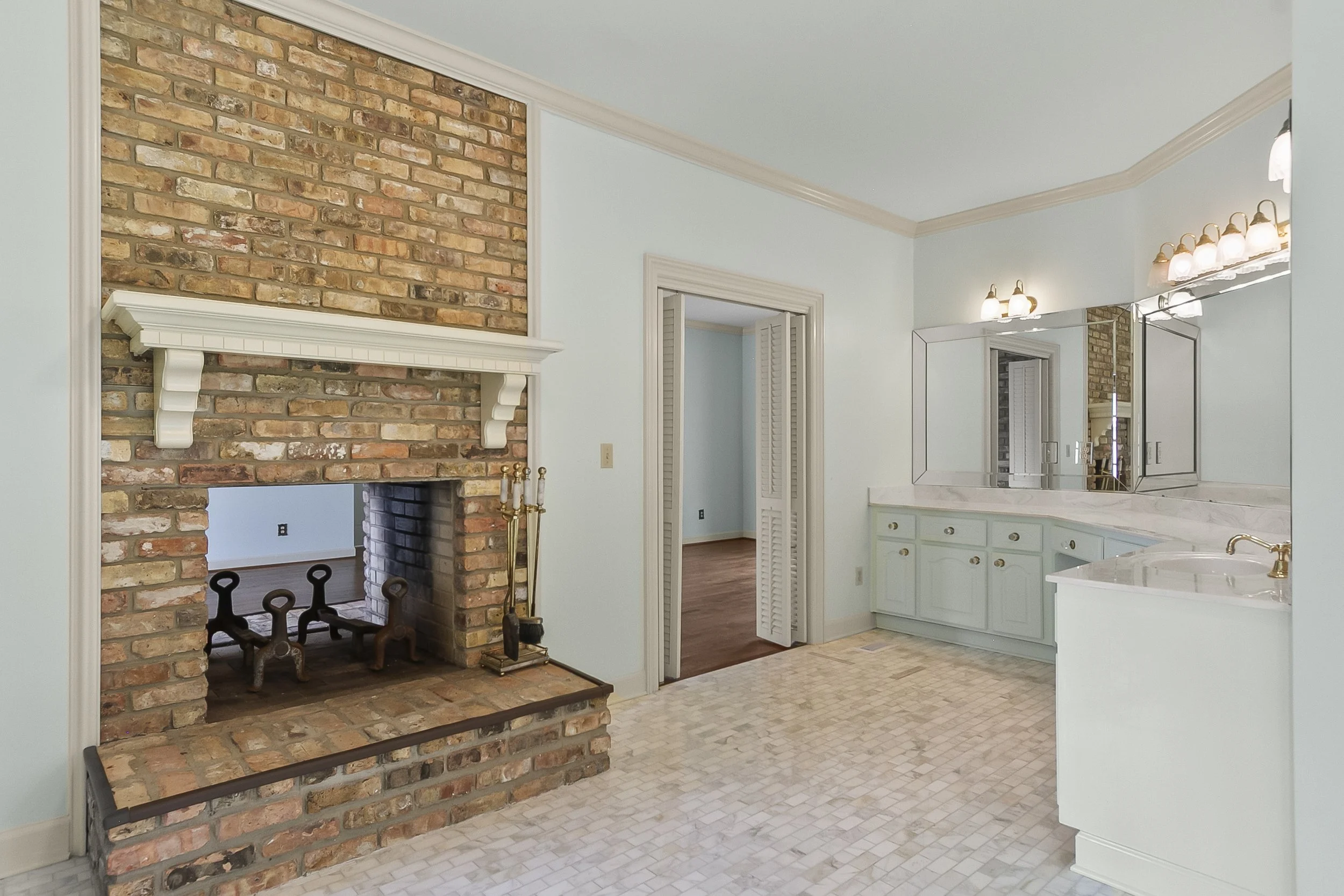 Empty bathroom with brick fireplace, light blue walls, white tiled floor, and a vanity with multiple mirrors and light fixtures.