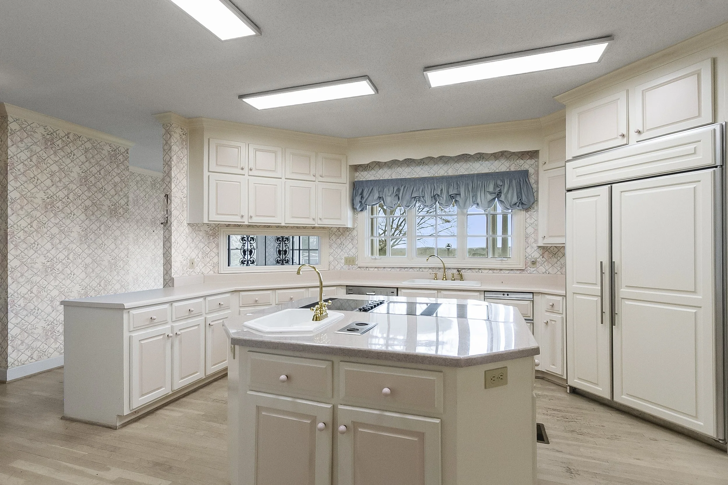 Bright white kitchen with cabinetry, a rounded island with a sink, and large windows with blue curtains.