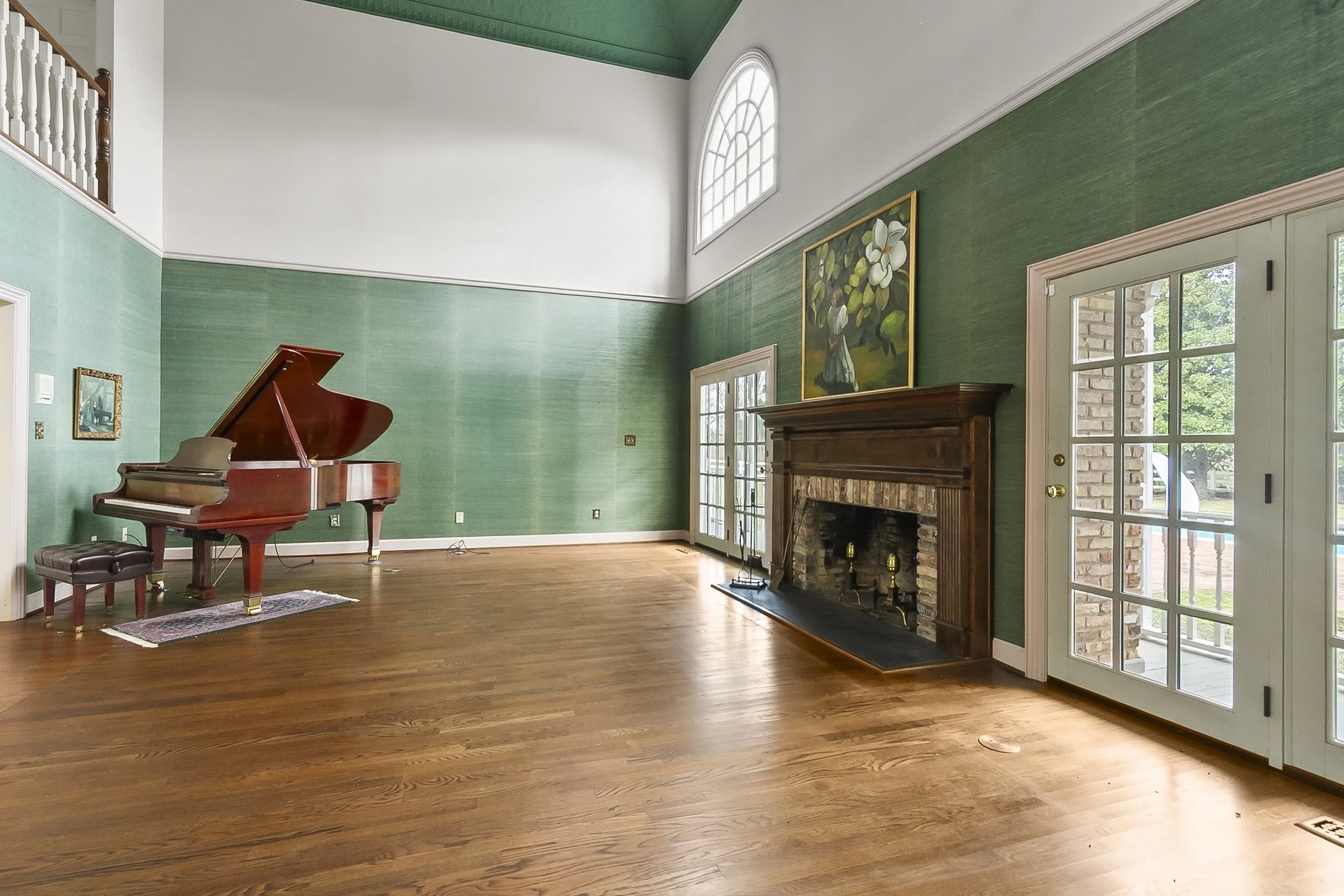 A spacious living room with hardwood floors, green textured walls, a brick fireplace with wooden mantel, and large glass doors leading outside. A grand piano and small bench are positioned near the walls, with a large floral painting hanging above th