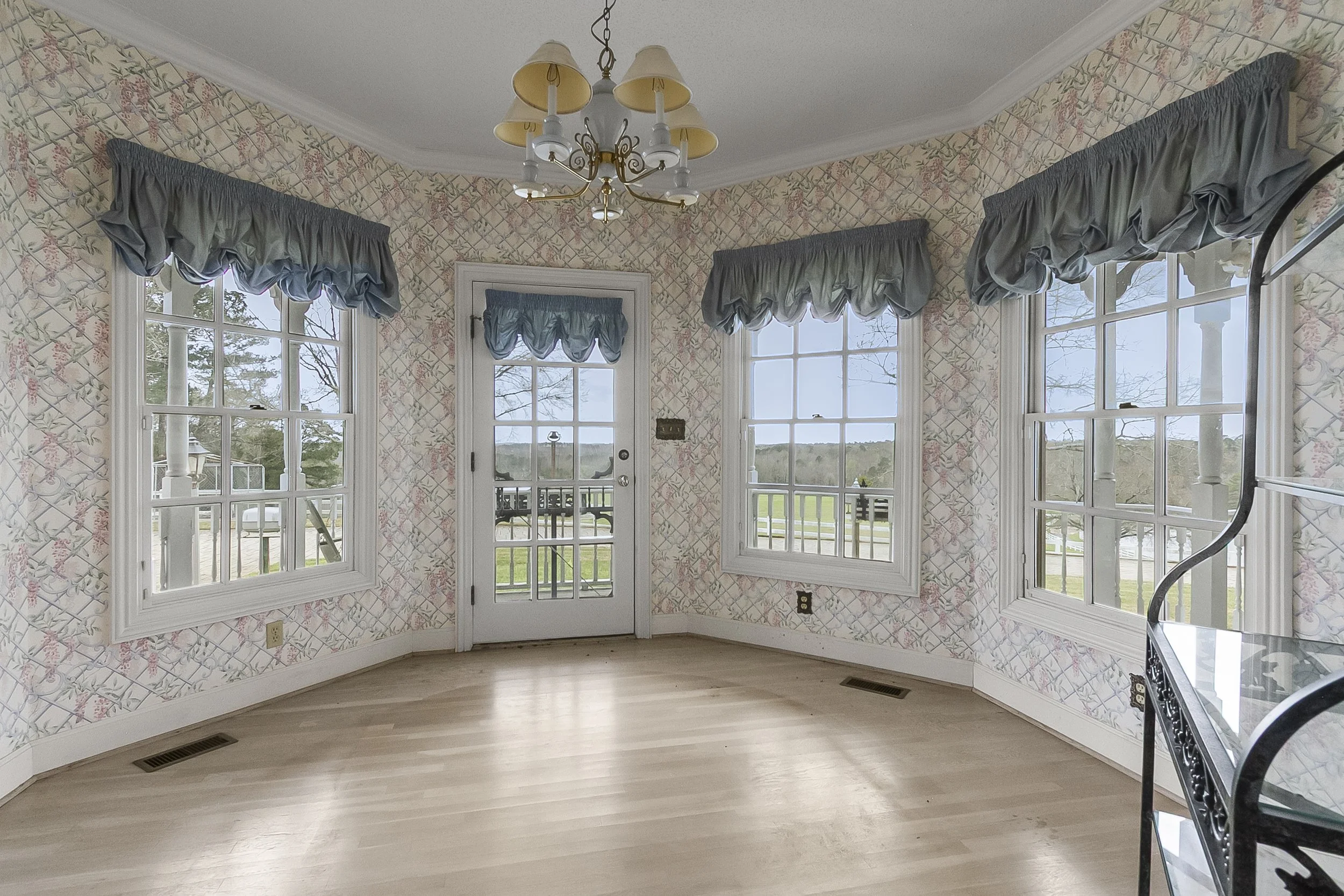 Empty room with floral wallpaper, hardwood floors, three large windows with blue valances, a glass door, a chandelier, and a metal stand in the corner.