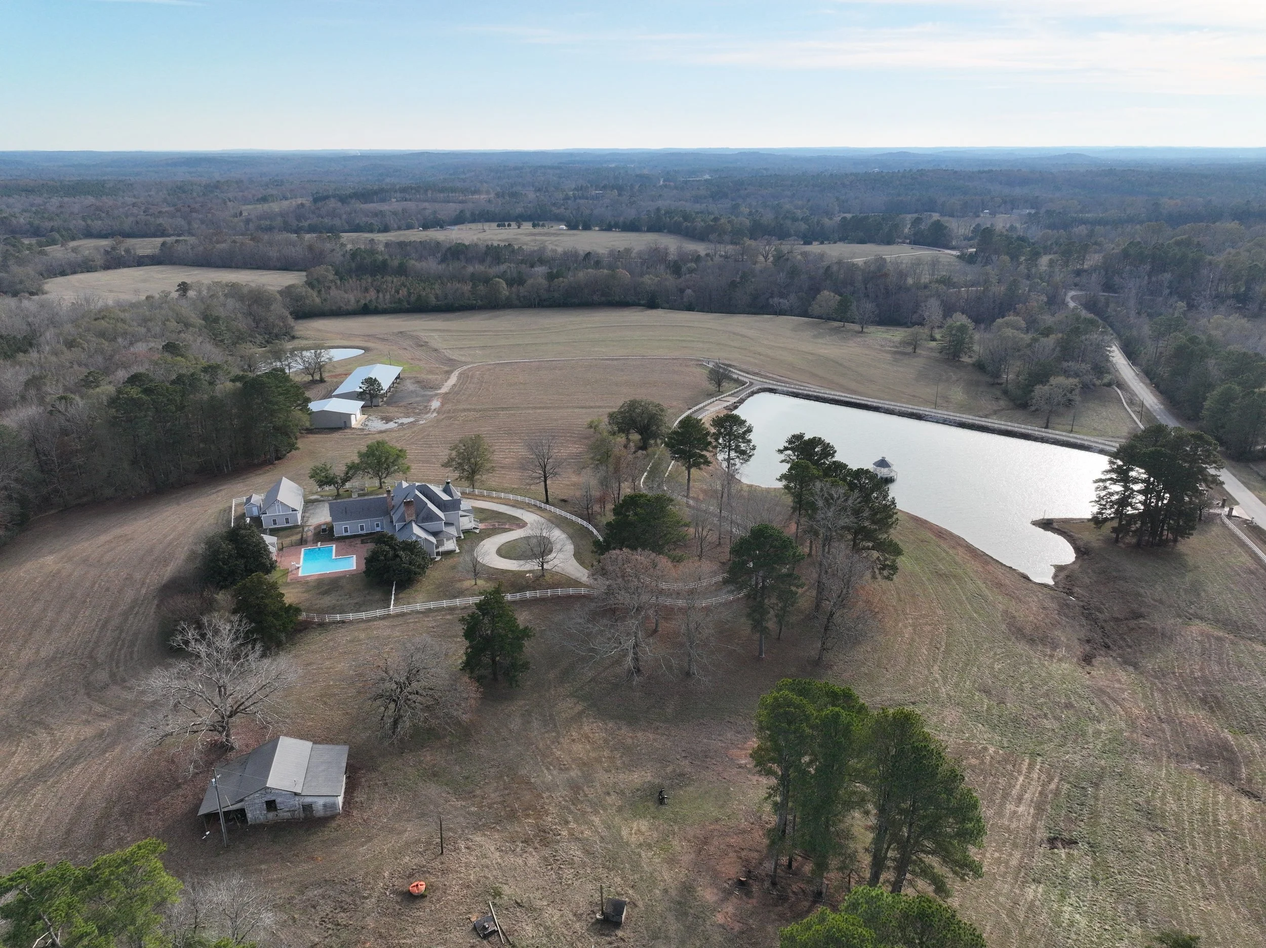Aerial view of a rural property with a large house, a swimming pool, a pond, surrounding trees, and open farmland extending towards the horizon.