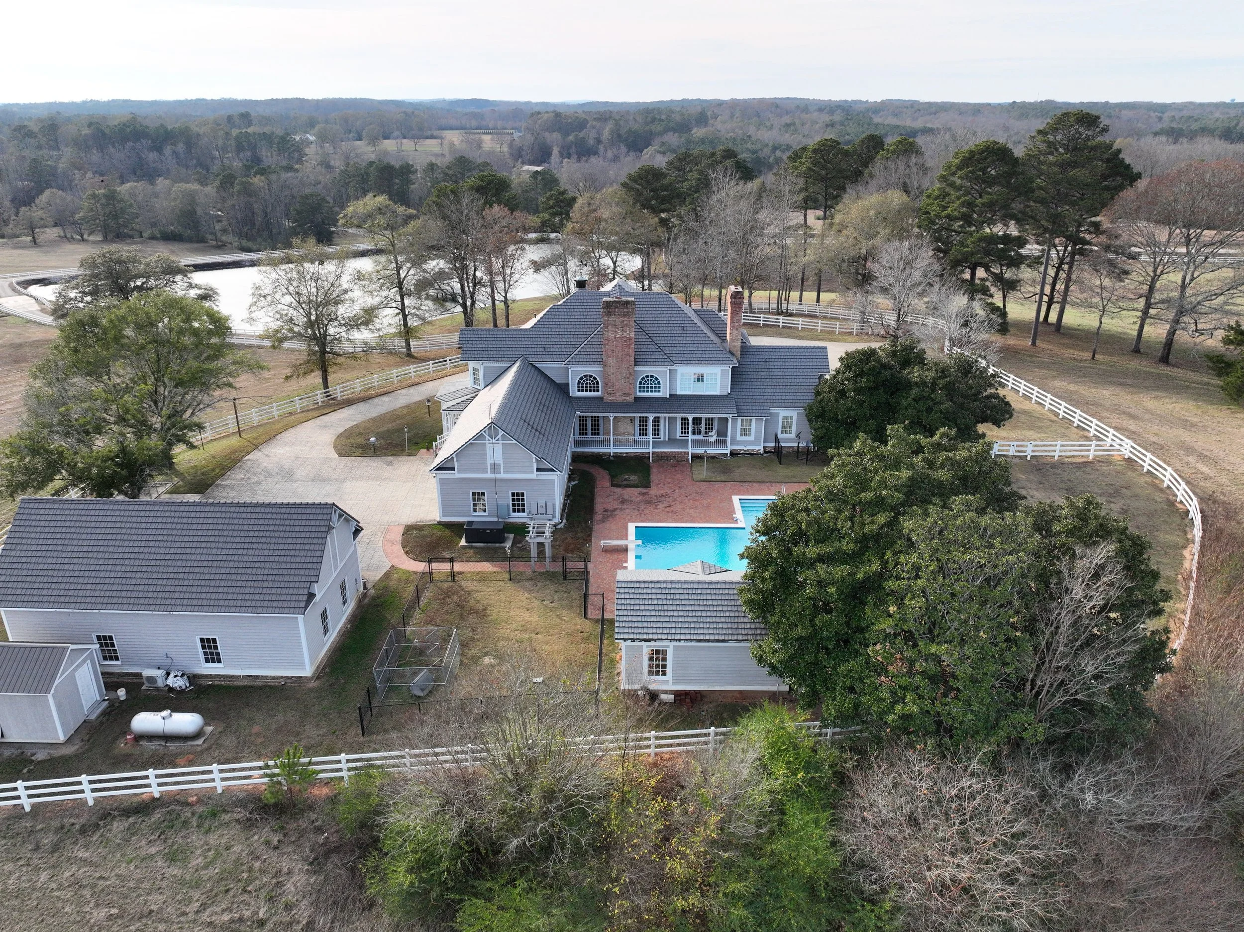 A large white house with a gray roof, a detached garage, and a swimming pool in the backyard surrounded by a white fence. The house is set on a spacious property with trees and open land, and there is a pond or small lake in the background.