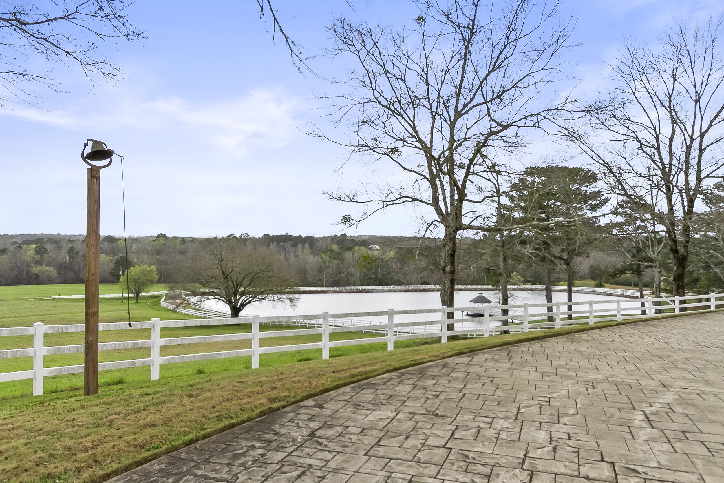 A serene outdoor scene with a paved walkway, white wooden fence, leafless trees, a pond, and a church with a steeple in the distance, under a partly cloudy sky.