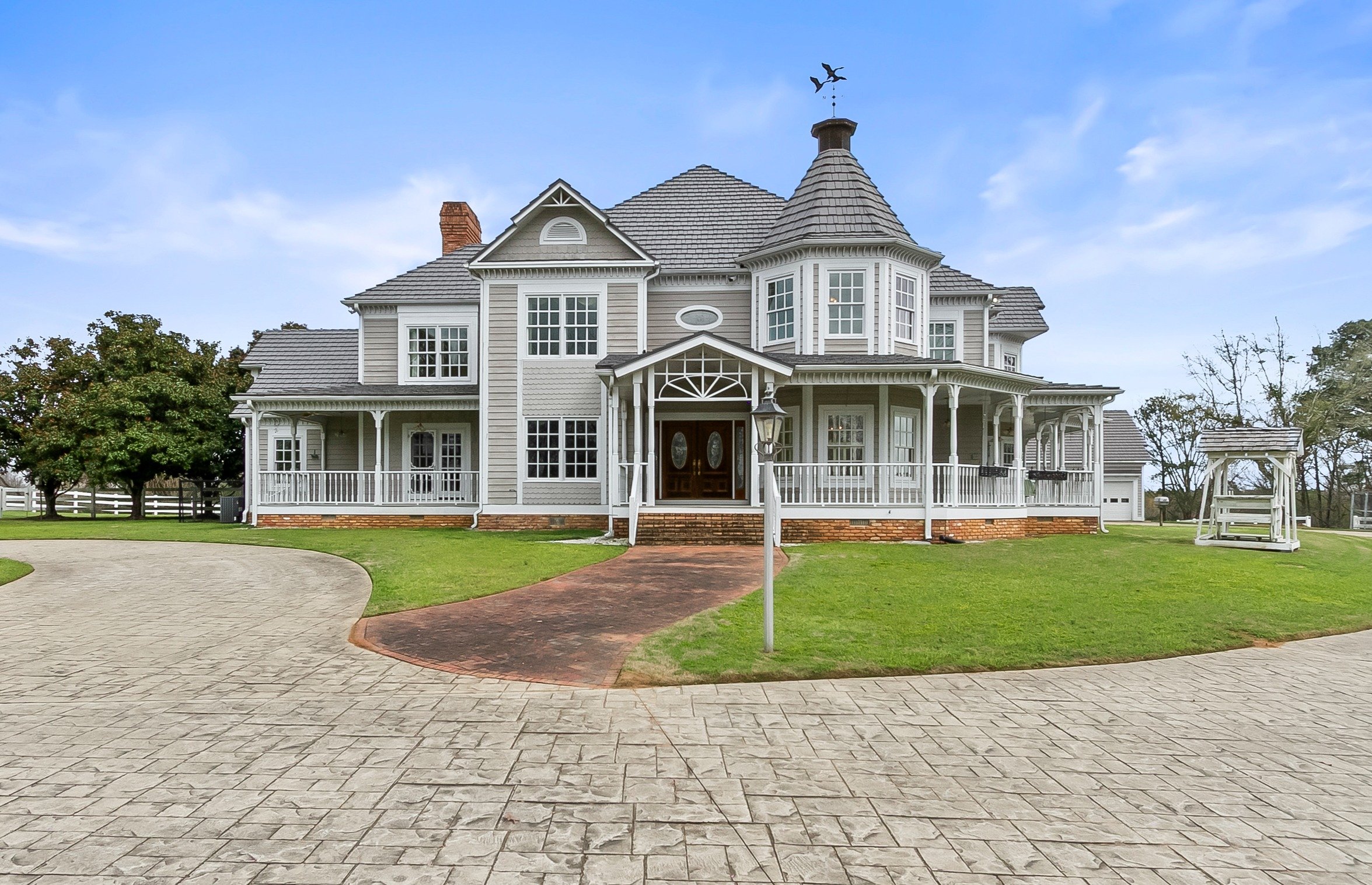 Large Victorian-style house with white siding, multiple gables, and a turret, surrounded by a landscaped lawn with trees and a curved driveway.