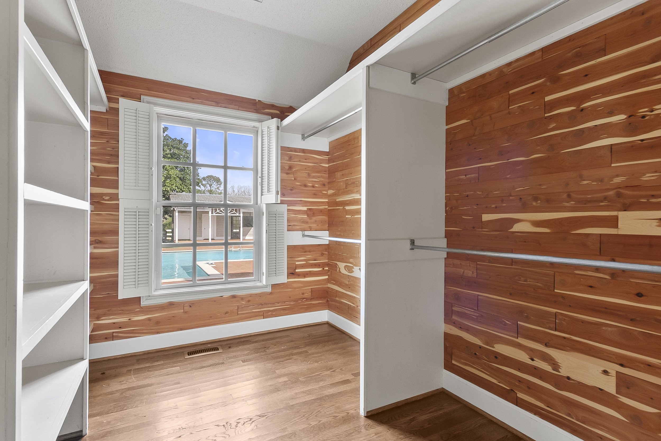 Empty walk-in closet with wooden walls, a large window with white shutters, a white shelf unit on the left, and a rod for hanging clothes.