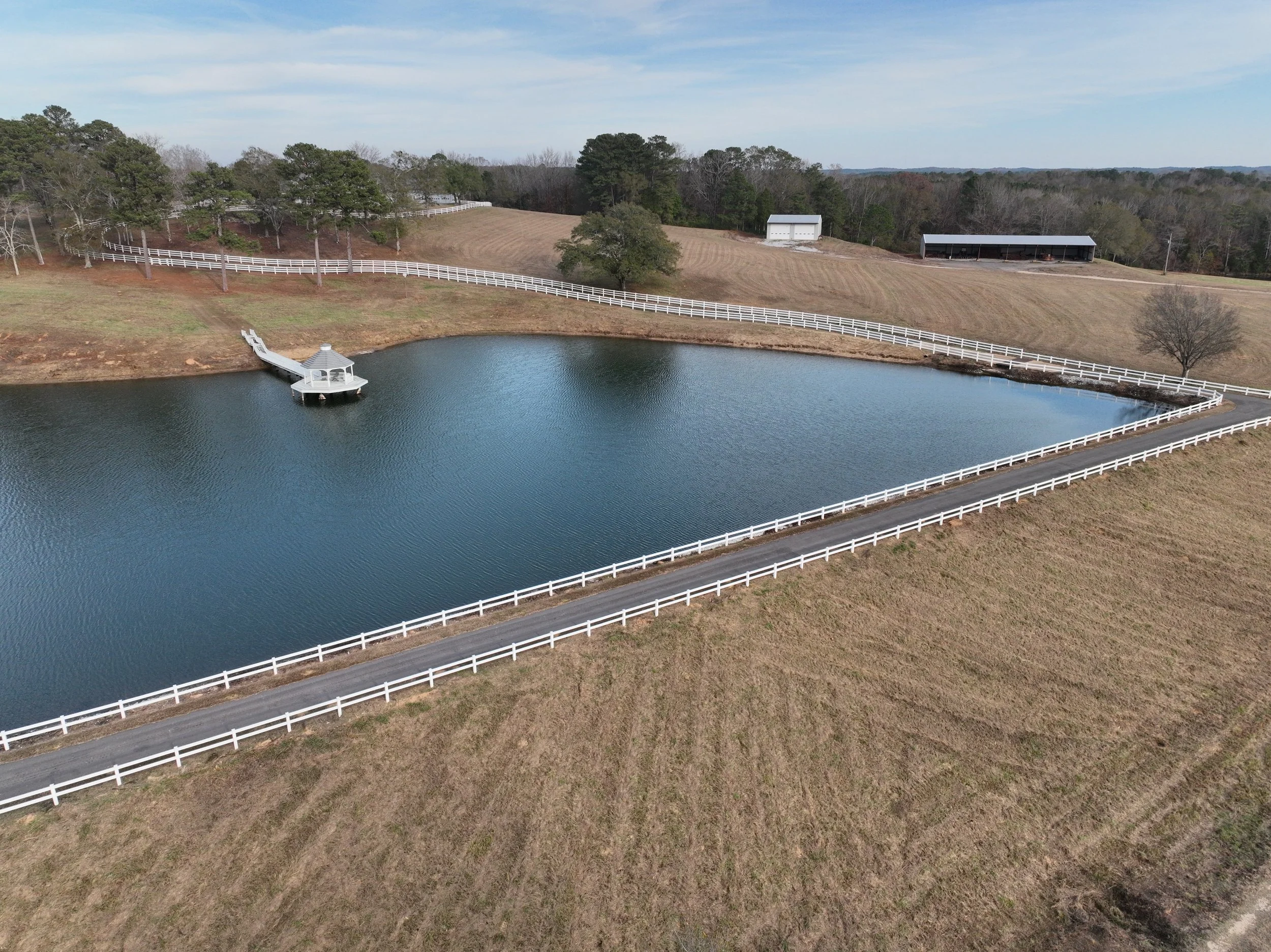 Aerial view of a small lake with a white gazebo and dock, surrounded by a white fence and farmland with trees and farm buildings in the background.