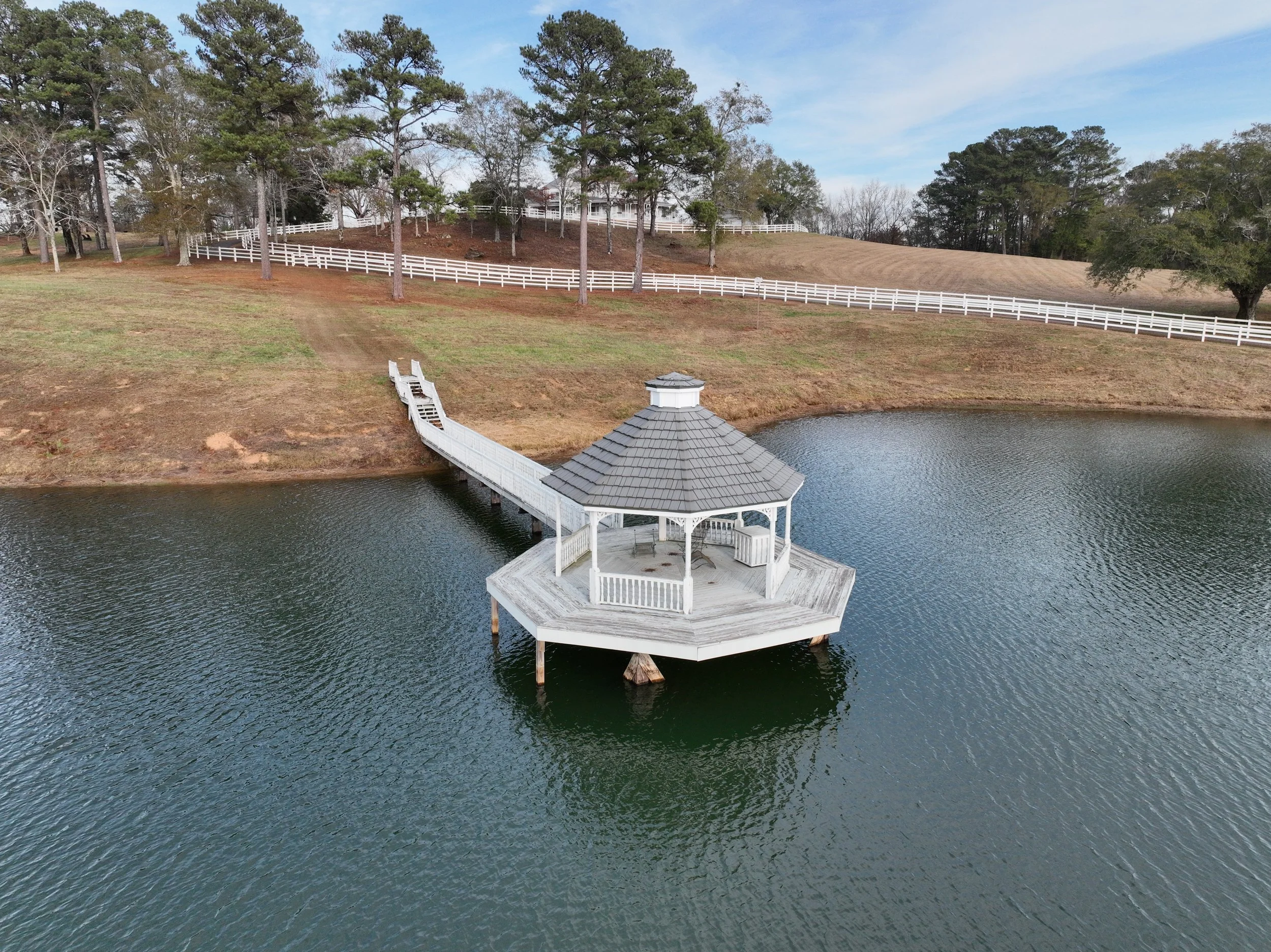 A white gazebo on a dock extending into a lake, with a wooden ramp leading to the grassy shore. Fenced land and trees are in the background under a partly cloudy sky.