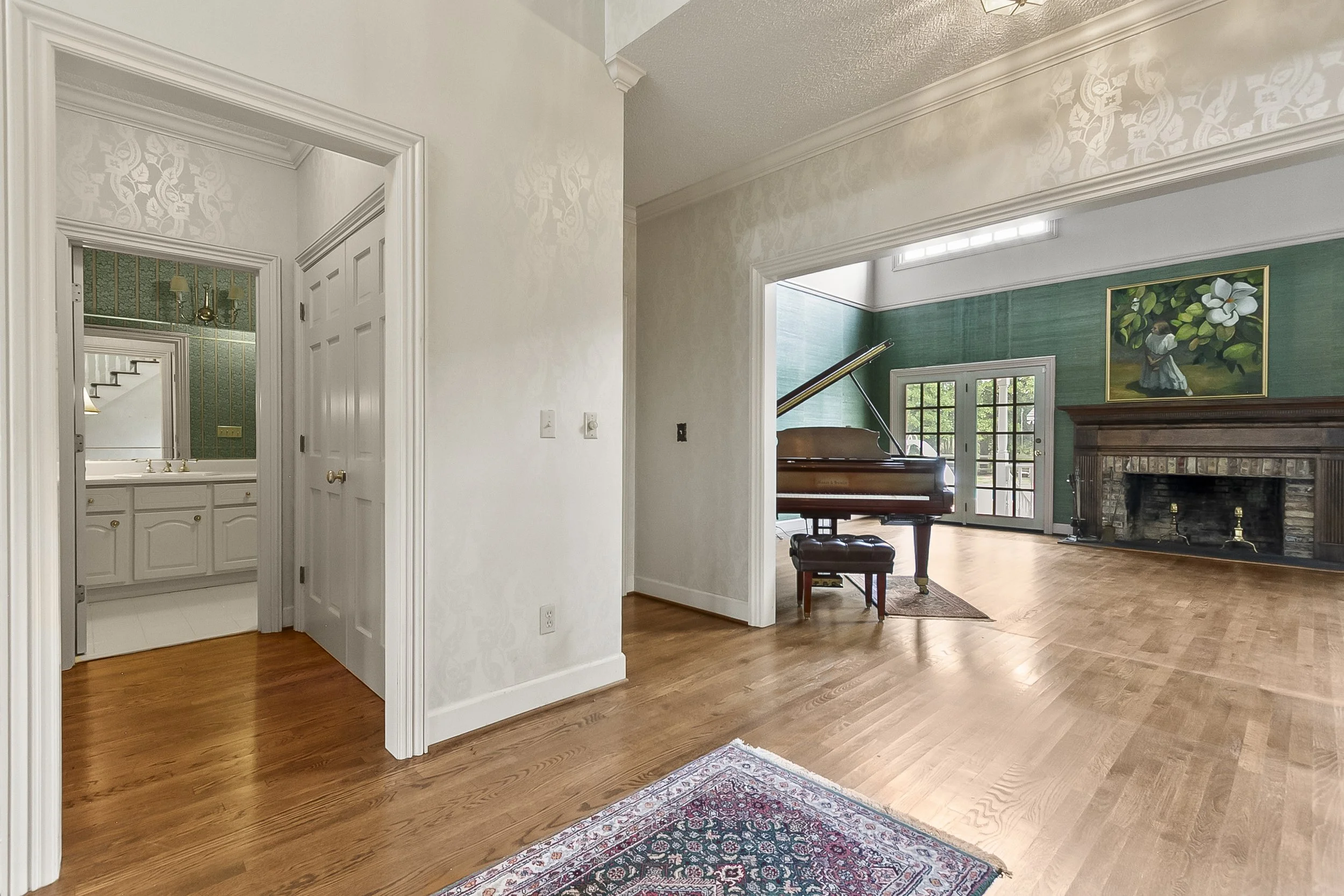 Interior view of a living room with a grand piano, a large fireplace, and French doors leading outside. Part of a bathroom with a white vanity and gold fixtures is visible through an open doorway. The room has hardwood floors and a decorative paintin