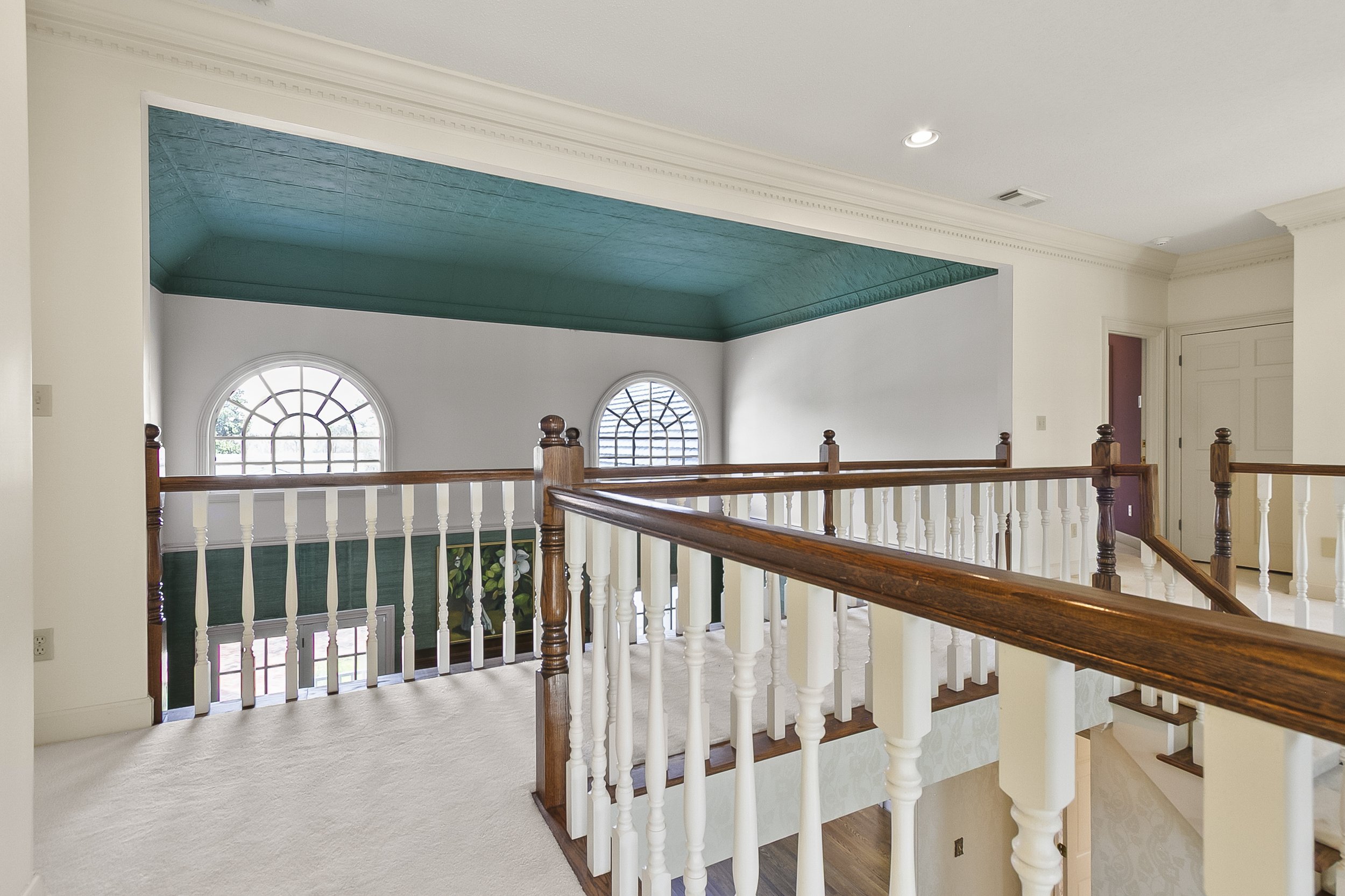 Interior view of a house landing with a wooden railing, white walls, turquoise ceiling, and two arched windows in the background.