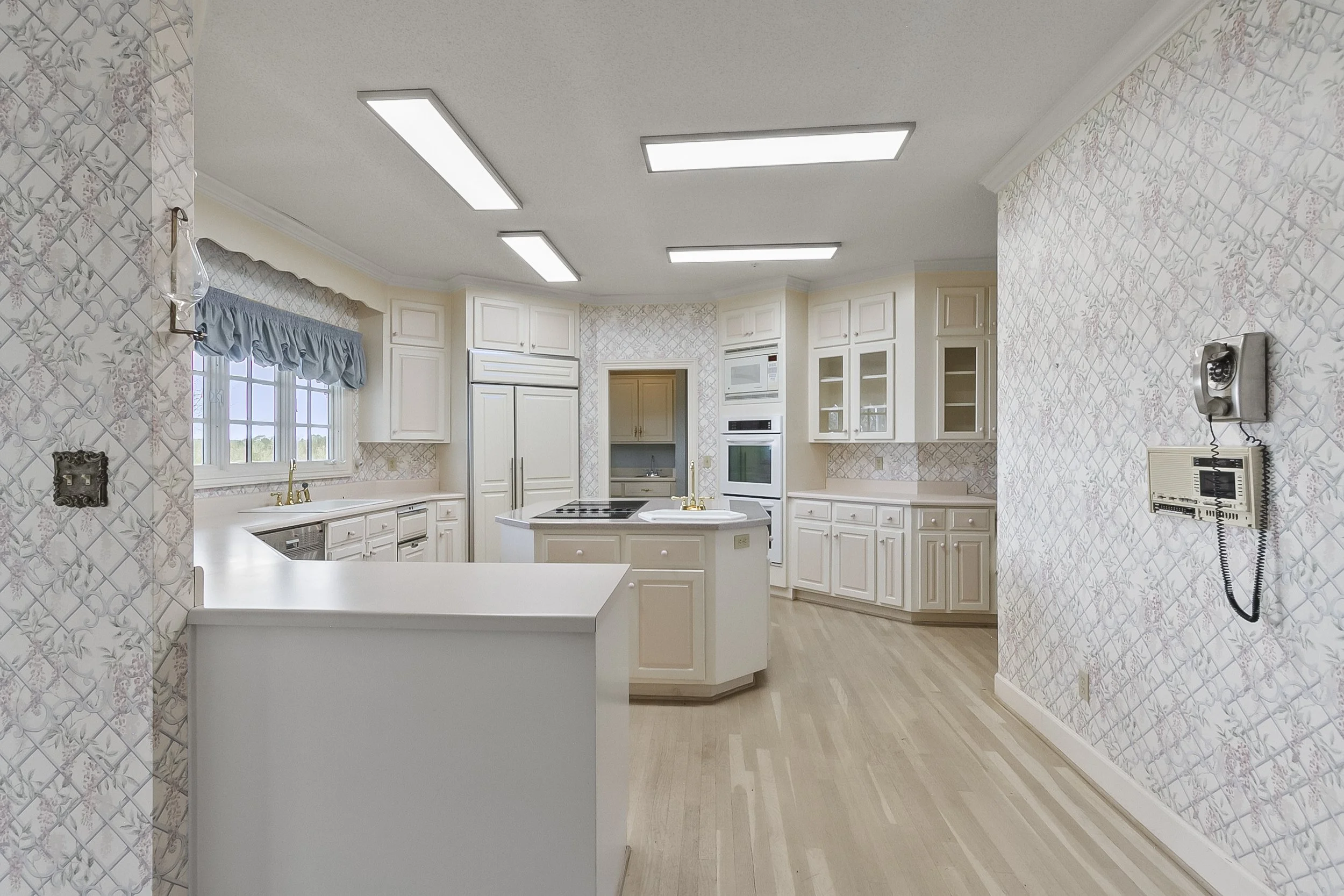 A bright, spacious kitchen with white cabinets, a central island with a stove, and a window with a blue valance. There are built-in appliances, a wall-mounted rotary phone, and wallpaper with a floral pattern.